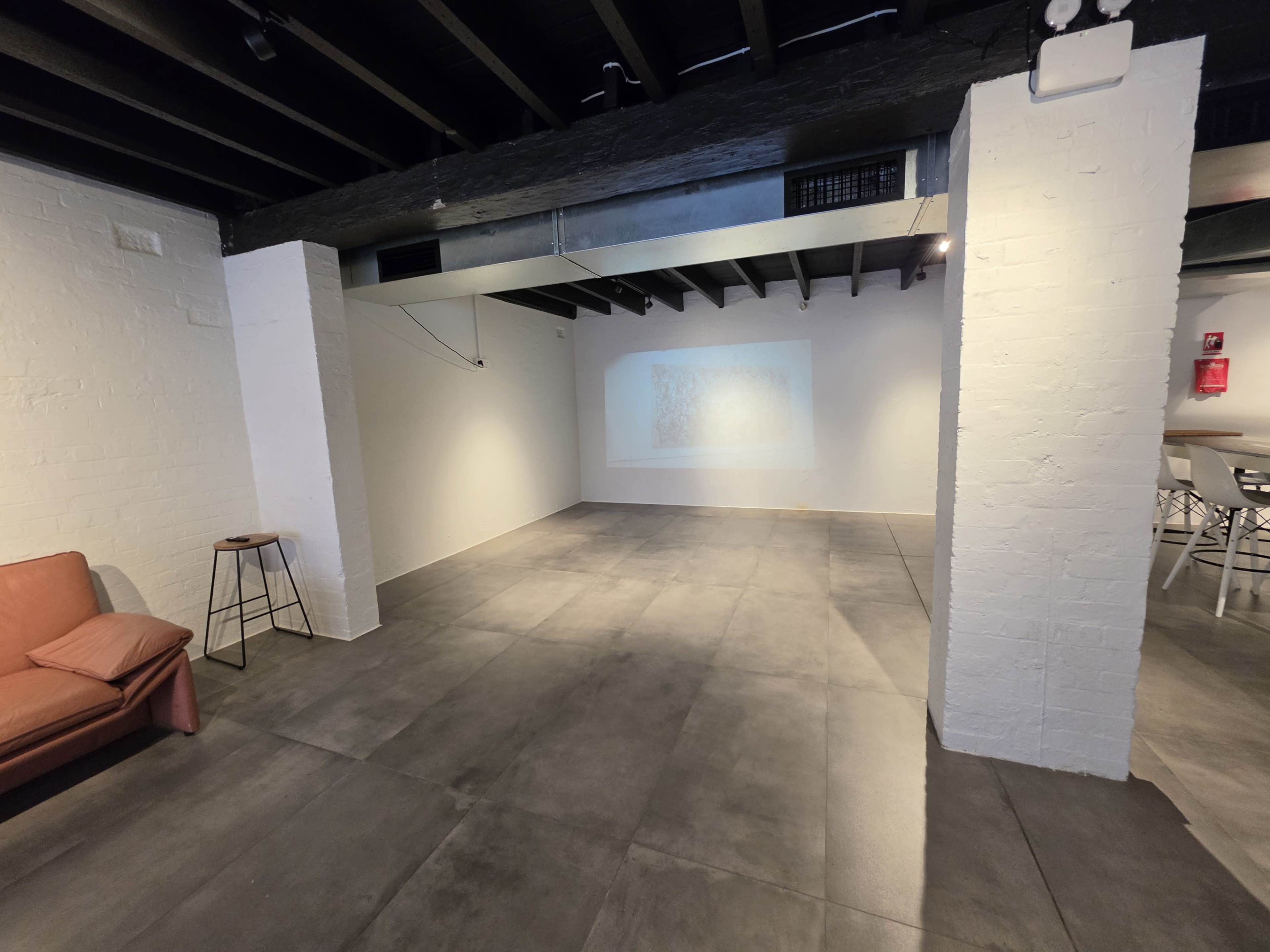 The image shows a spacious, minimalist room with white walls, a concrete floor, and a small sofa in one corner.