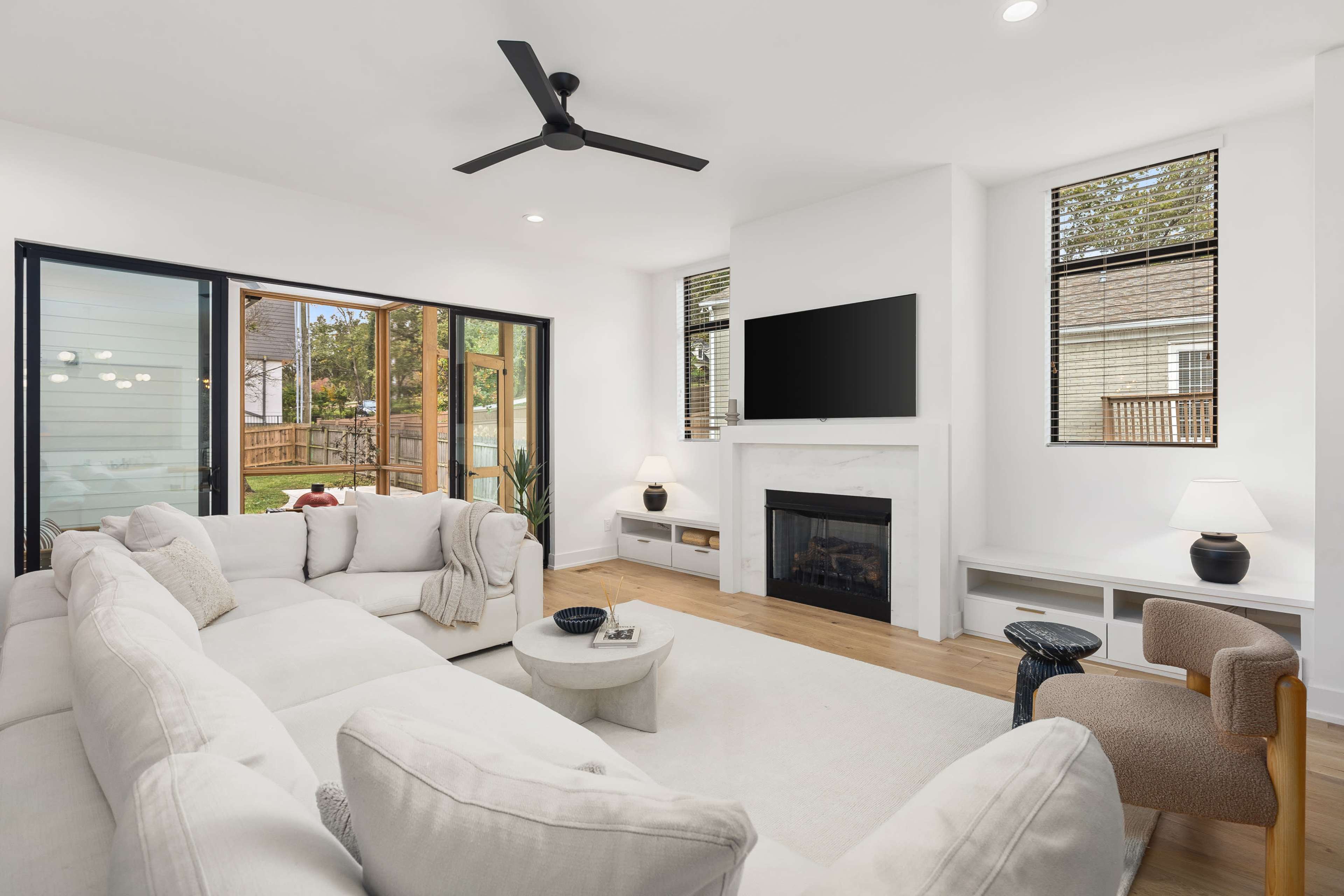 The living room features a light-colored sectional sofa, a round coffee table, a wall-mounted television, and a fireplace, with large windows providing views of the outdoor space.