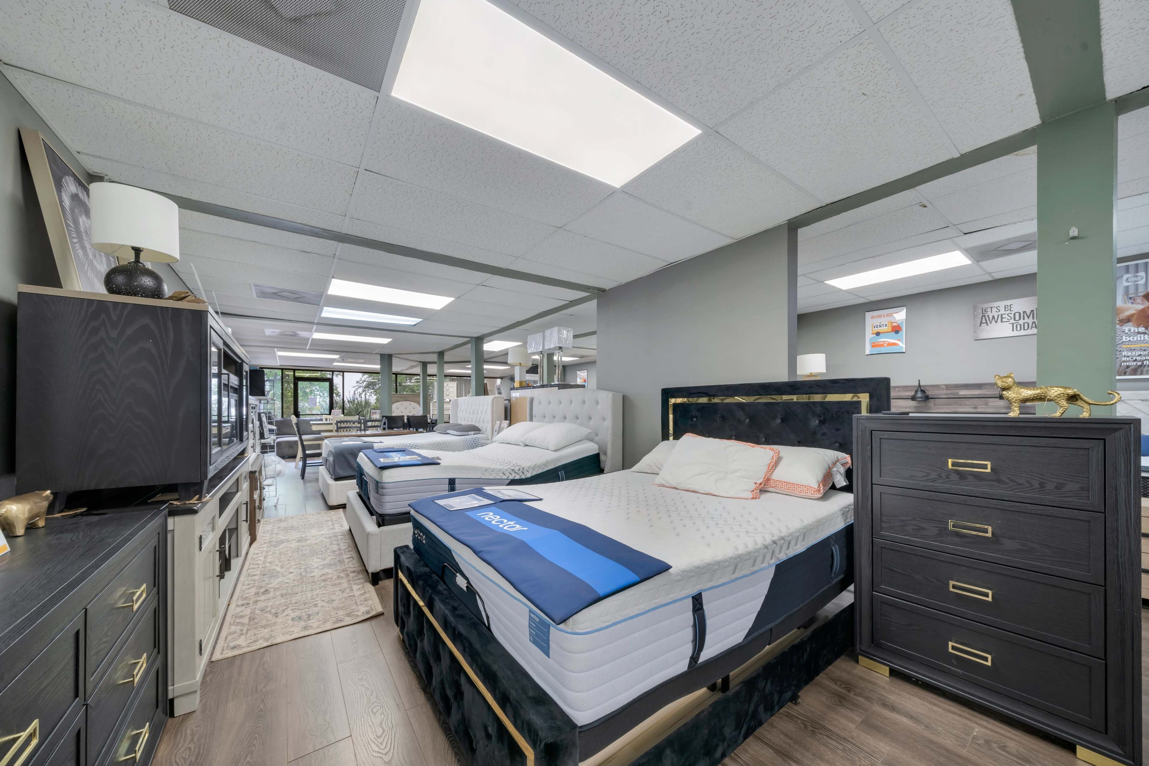 The image shows a showroom displaying several beds with mattresses, a dresser, and various other furniture pieces under bright lighting.