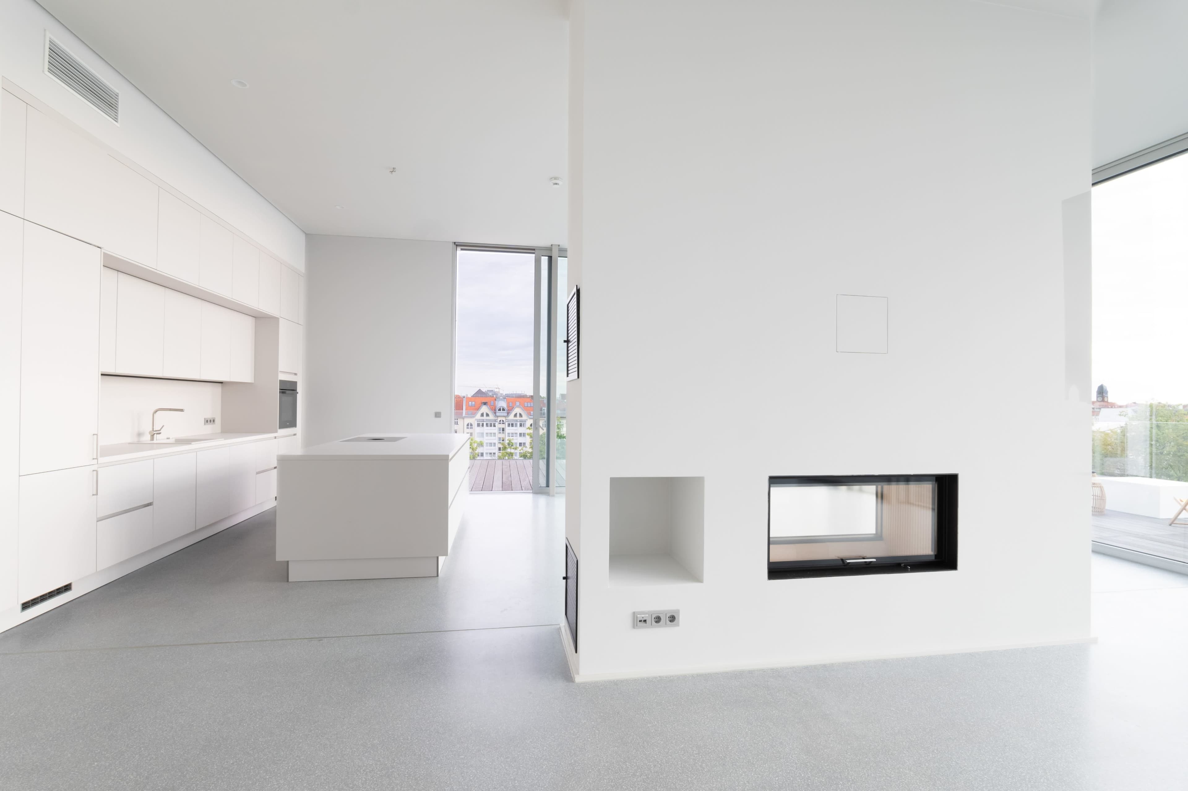 The image shows a modern, open-concept kitchen and living area with a sleek design, featuring minimalist cabinetry and a built-in fireplace.