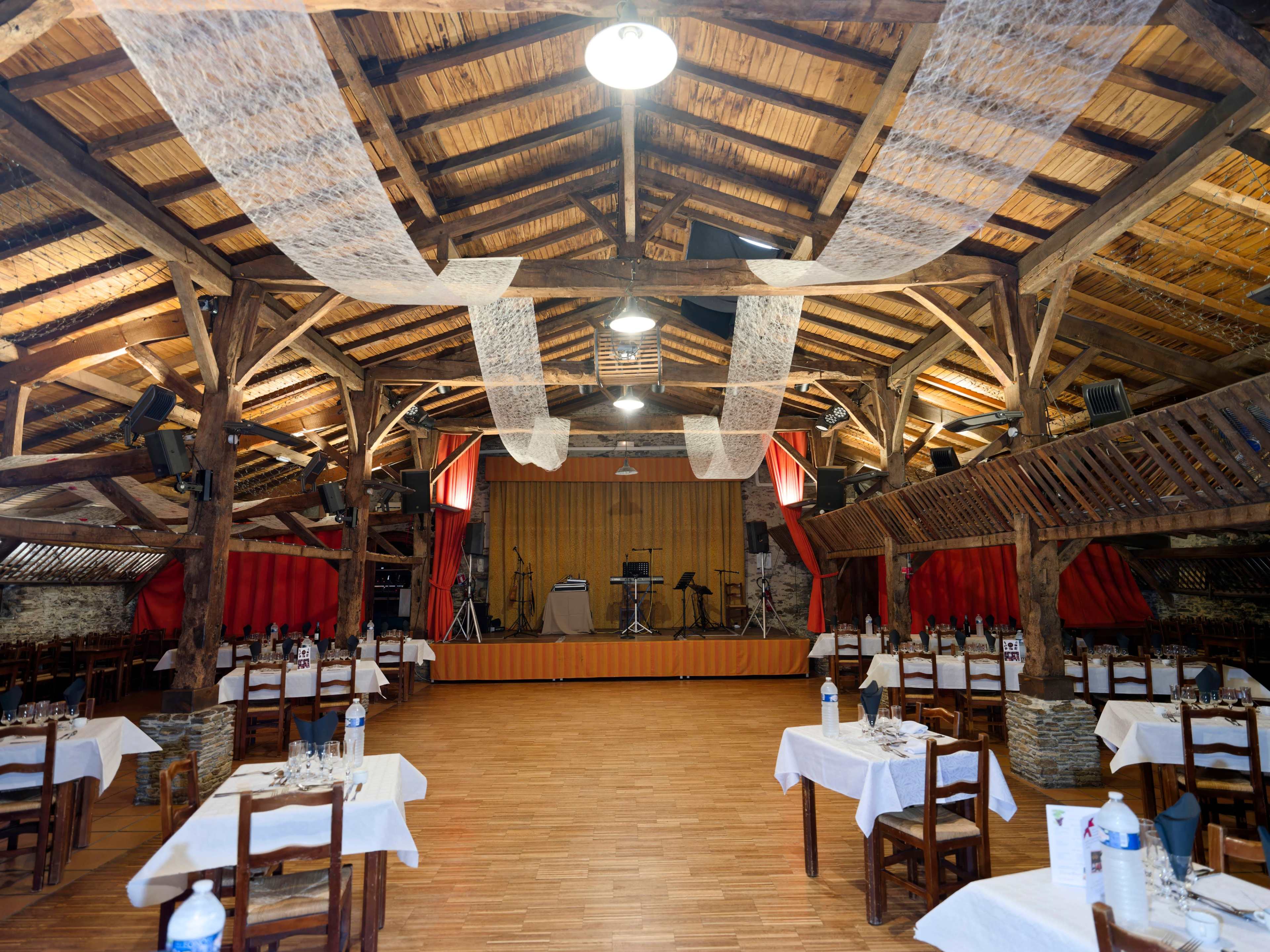 The image shows a spacious restaurant interior with wooden beams, tables set for dining, and a small stage at one end.