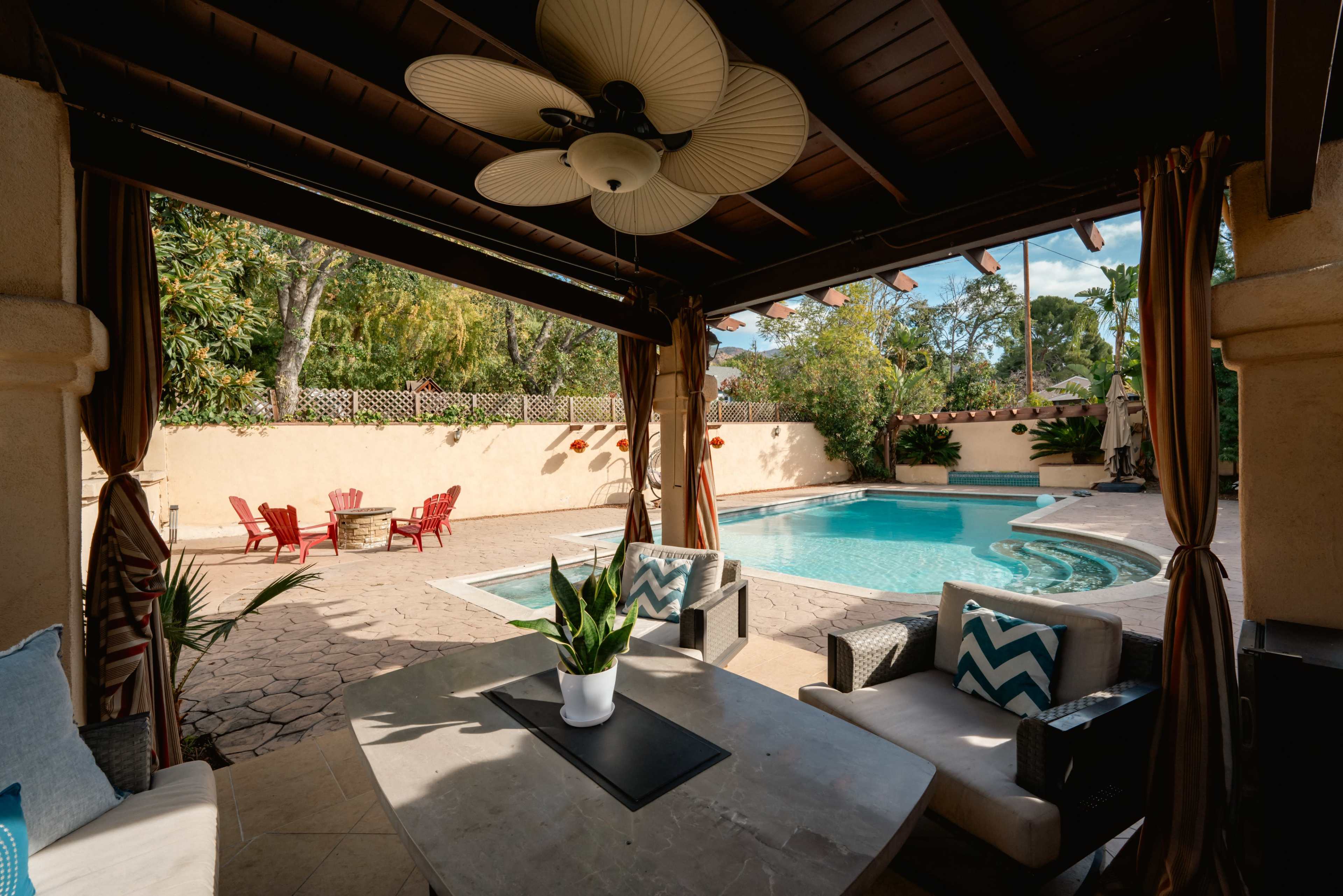 A shaded patio area features a table with chairs overlooking a swimming pool surrounded by landscaped greenery.