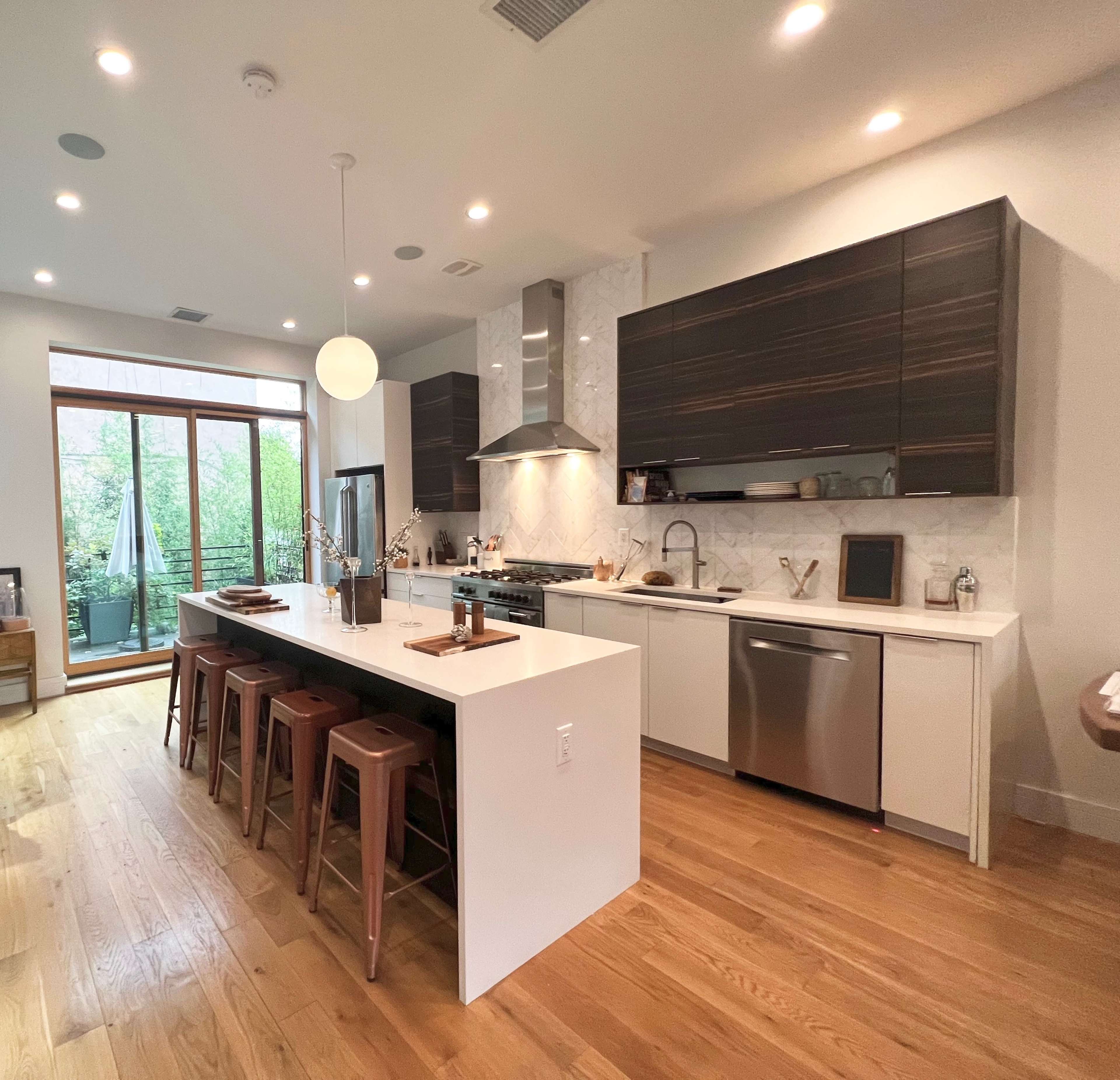 The image shows a modern kitchen featuring a large island with bar seating, stainless steel appliances, and ample natural light coming through large windows.