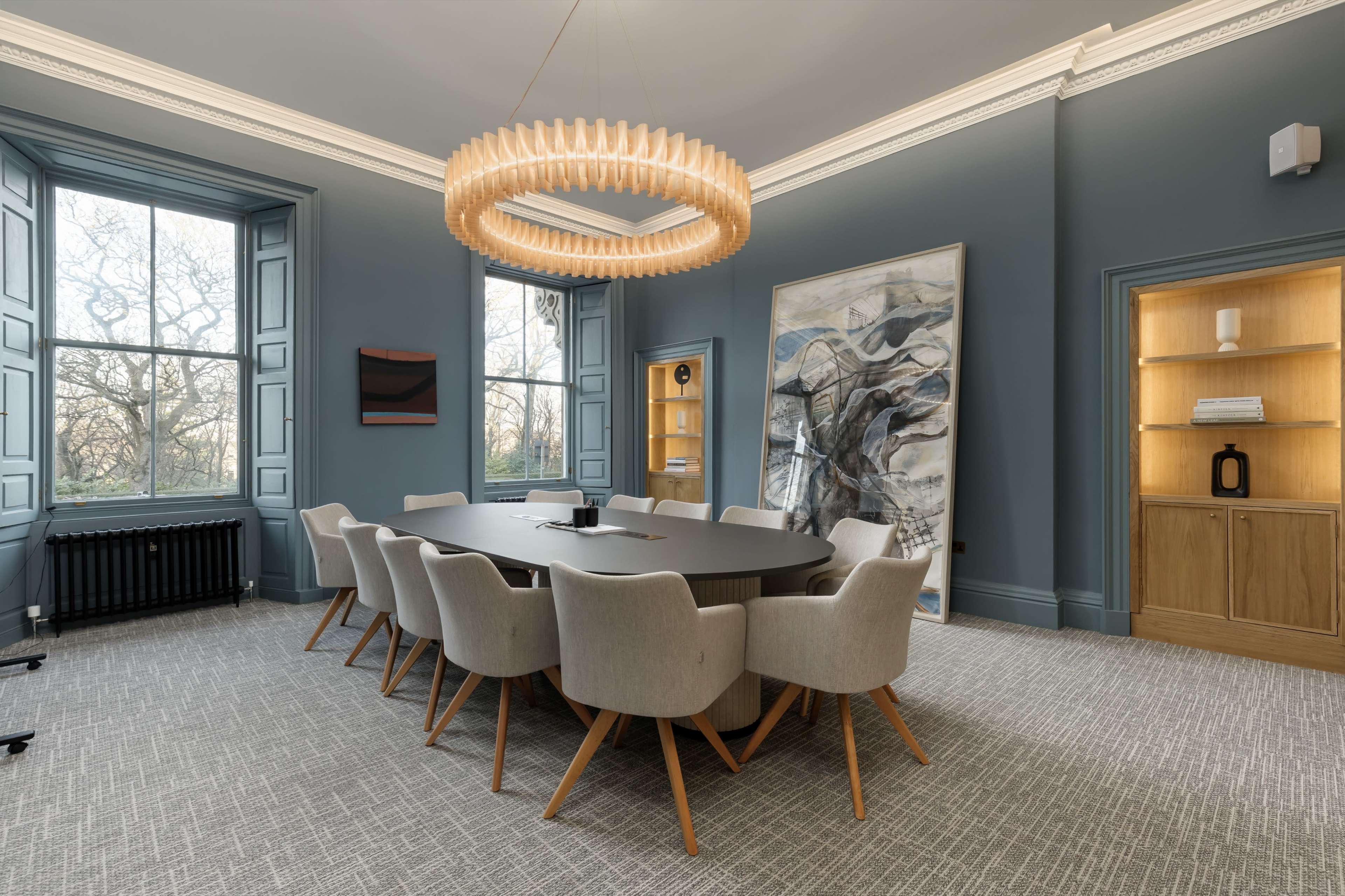 A modern conference room features a large oval table surrounded by upholstered chairs, illuminated by a distinctive circular chandelier, with large windows providing natural light.