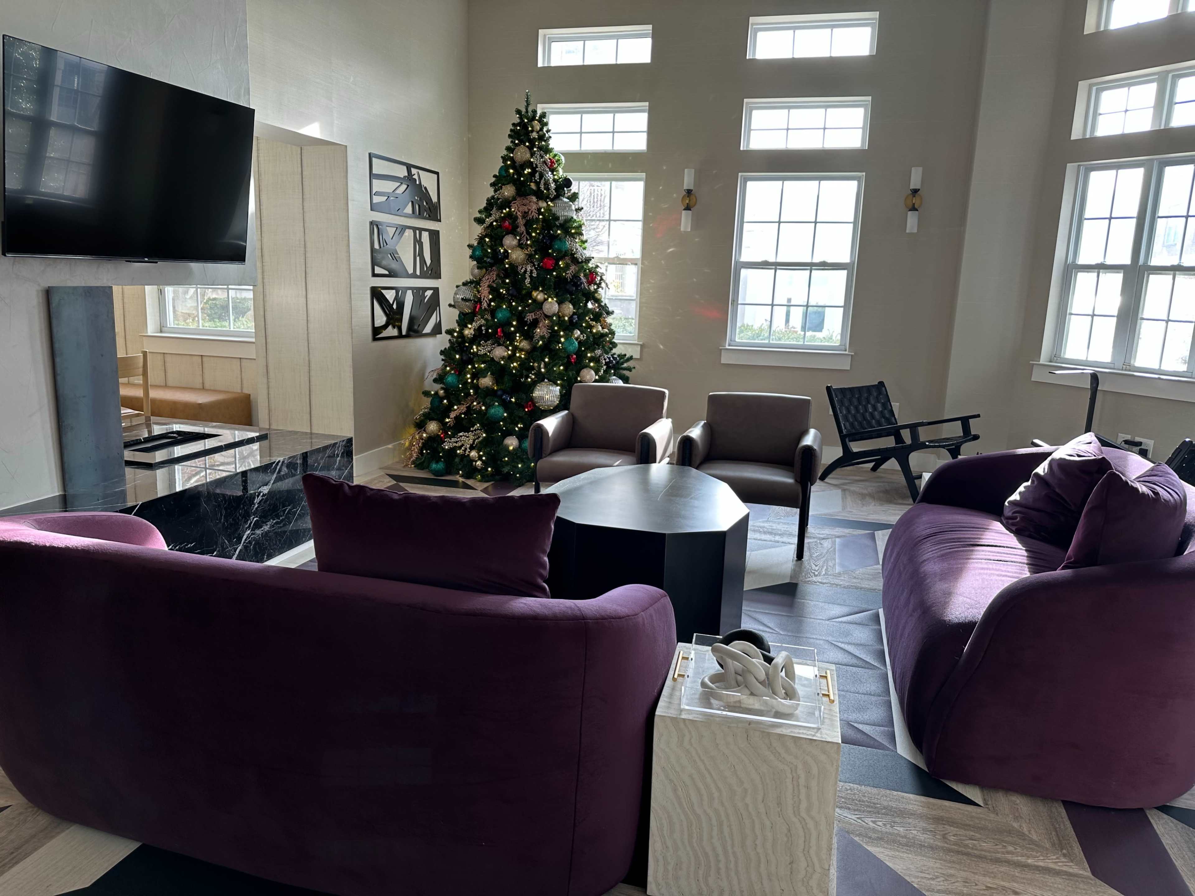 A cozy living area features a large Christmas tree surrounded by purple sofas, armchairs, and a circular coffee table.