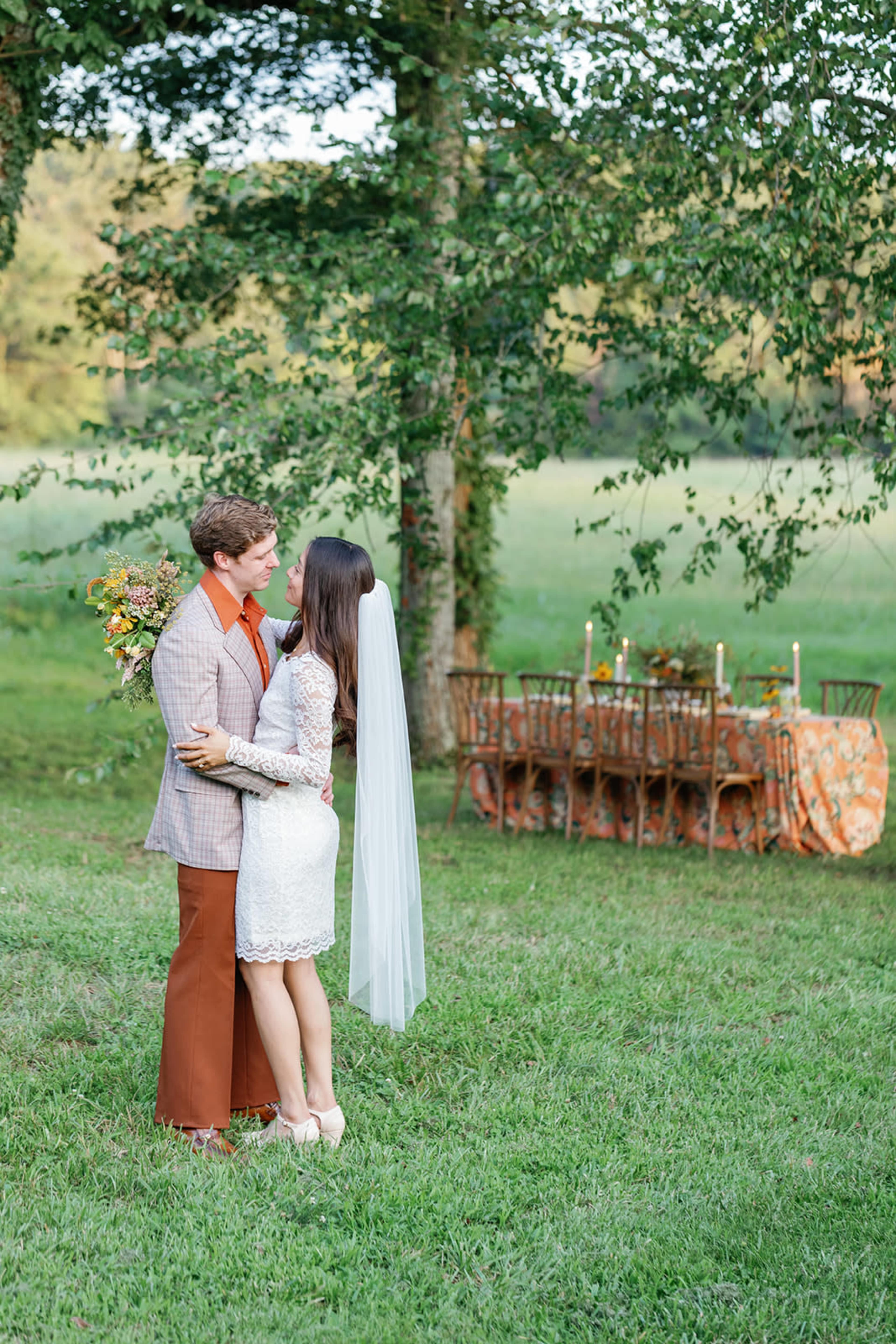 A couple embraces in a grassy field, surrounded by trees and a decorated table set for an outdoor celebration.