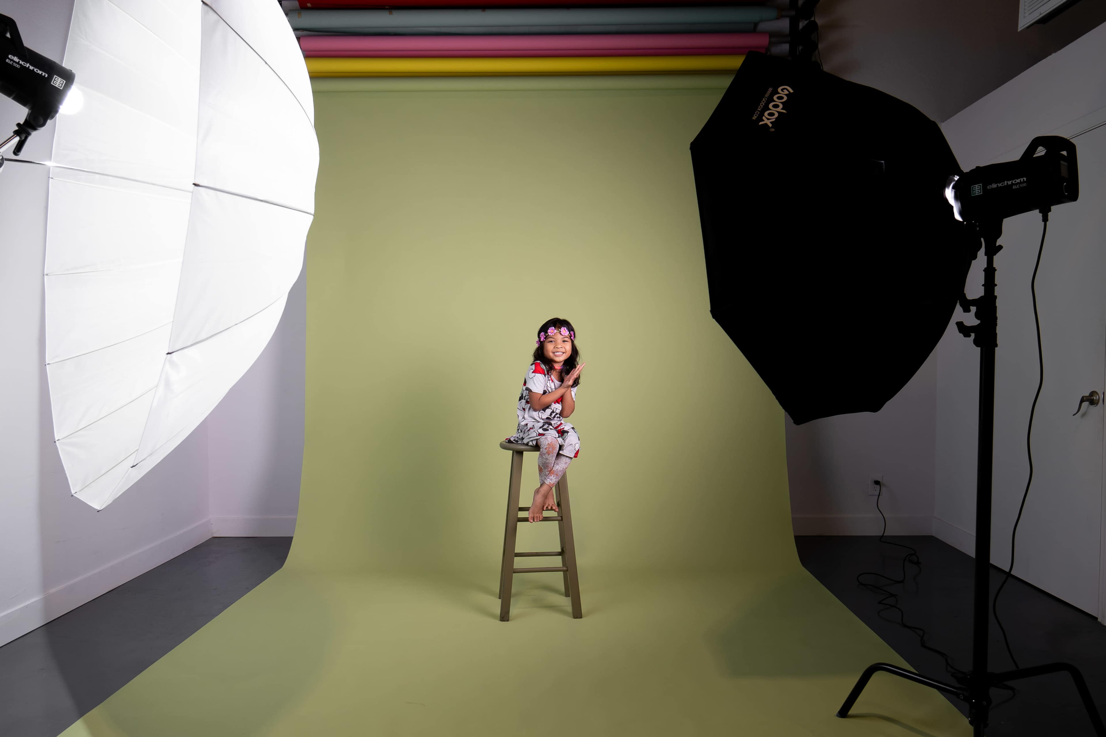 A child sits on a wooden stool in front of a green backdrop, illuminated by two large studio lights.