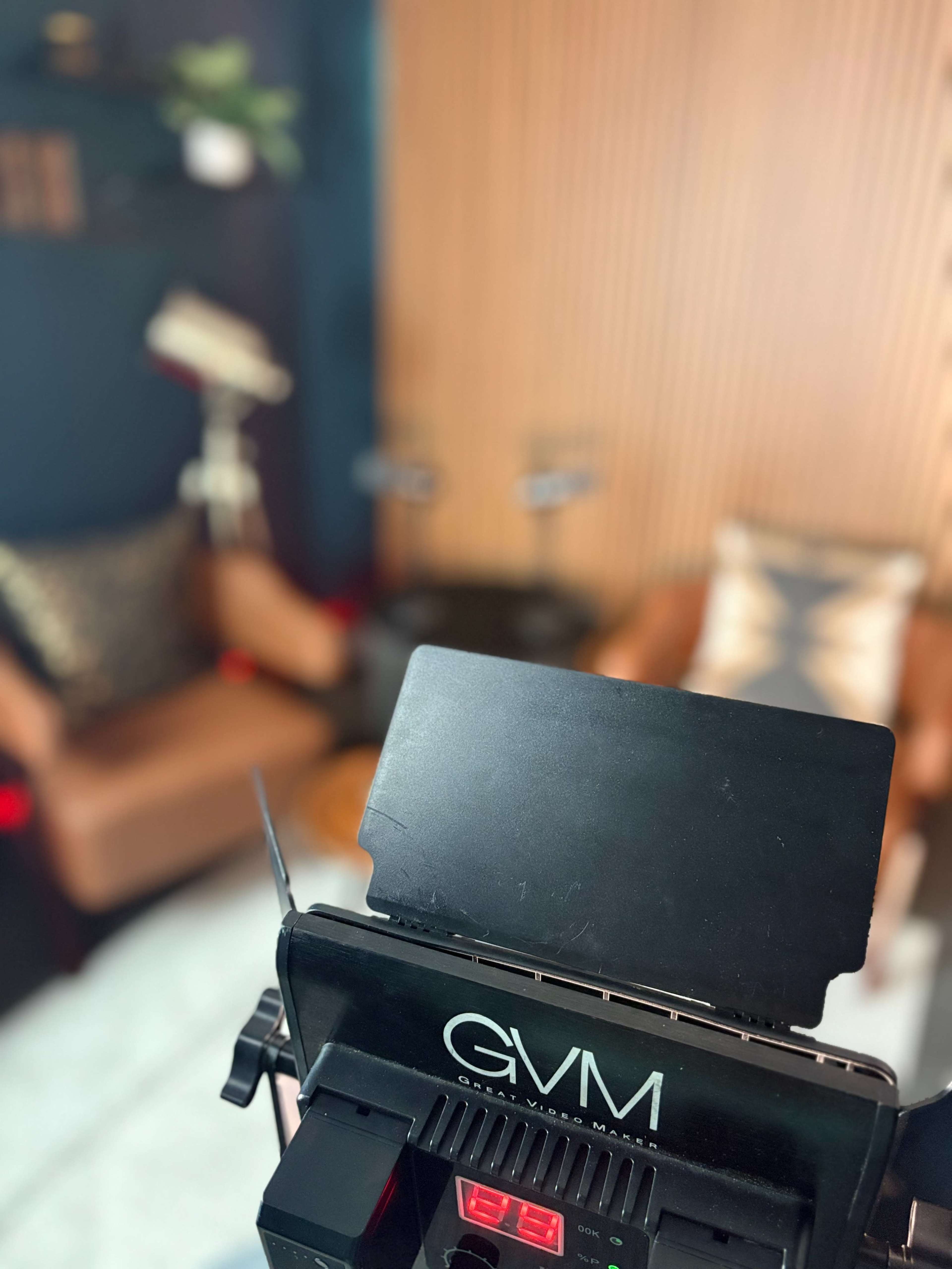 The image shows a video production setup with a GVM lighting panel in the foreground and a cozy seating area featuring two brown chairs in the background.