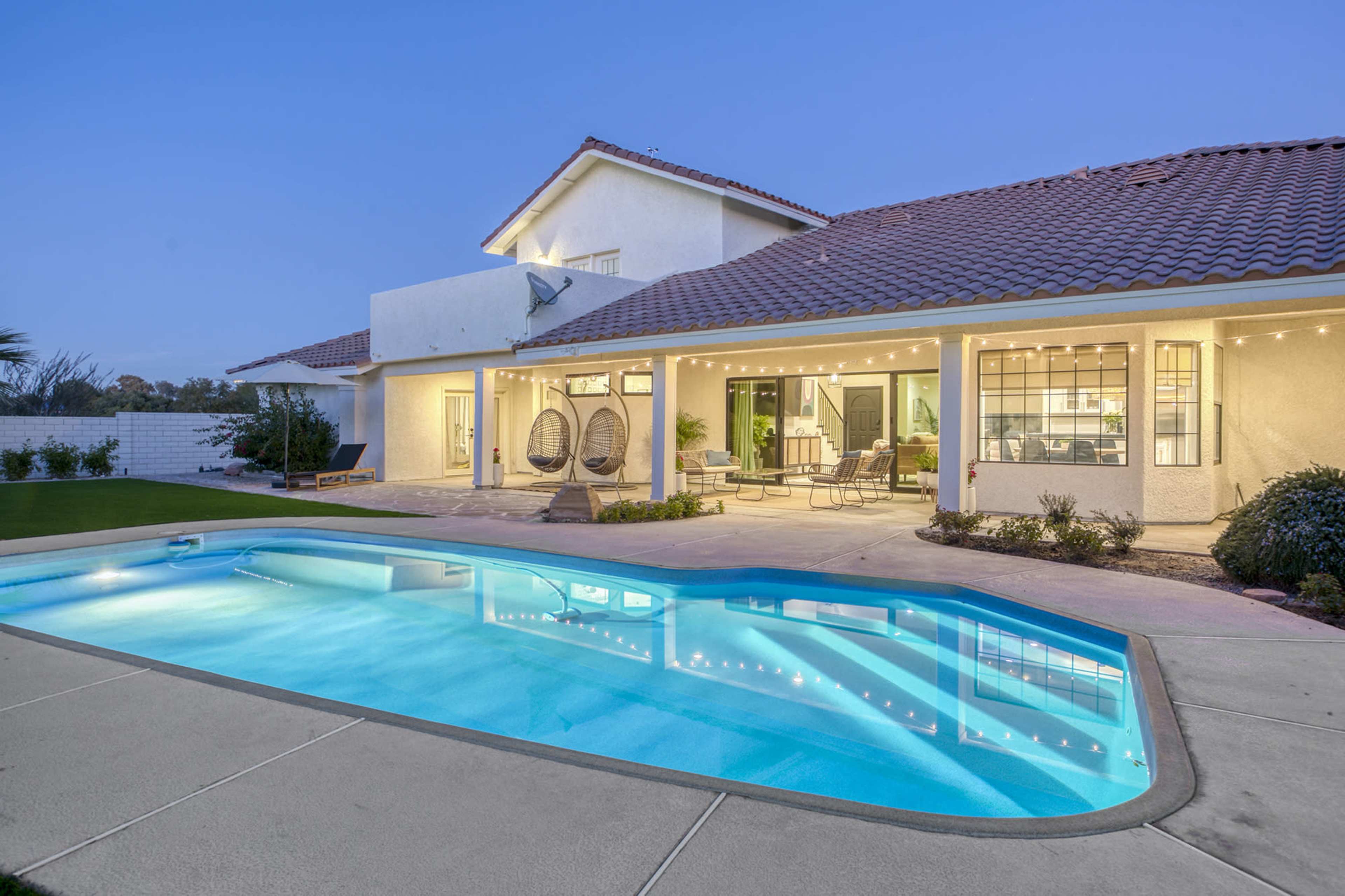 A two-story house features a pool in the backyard with hanging chairs and patio seating under string lights.