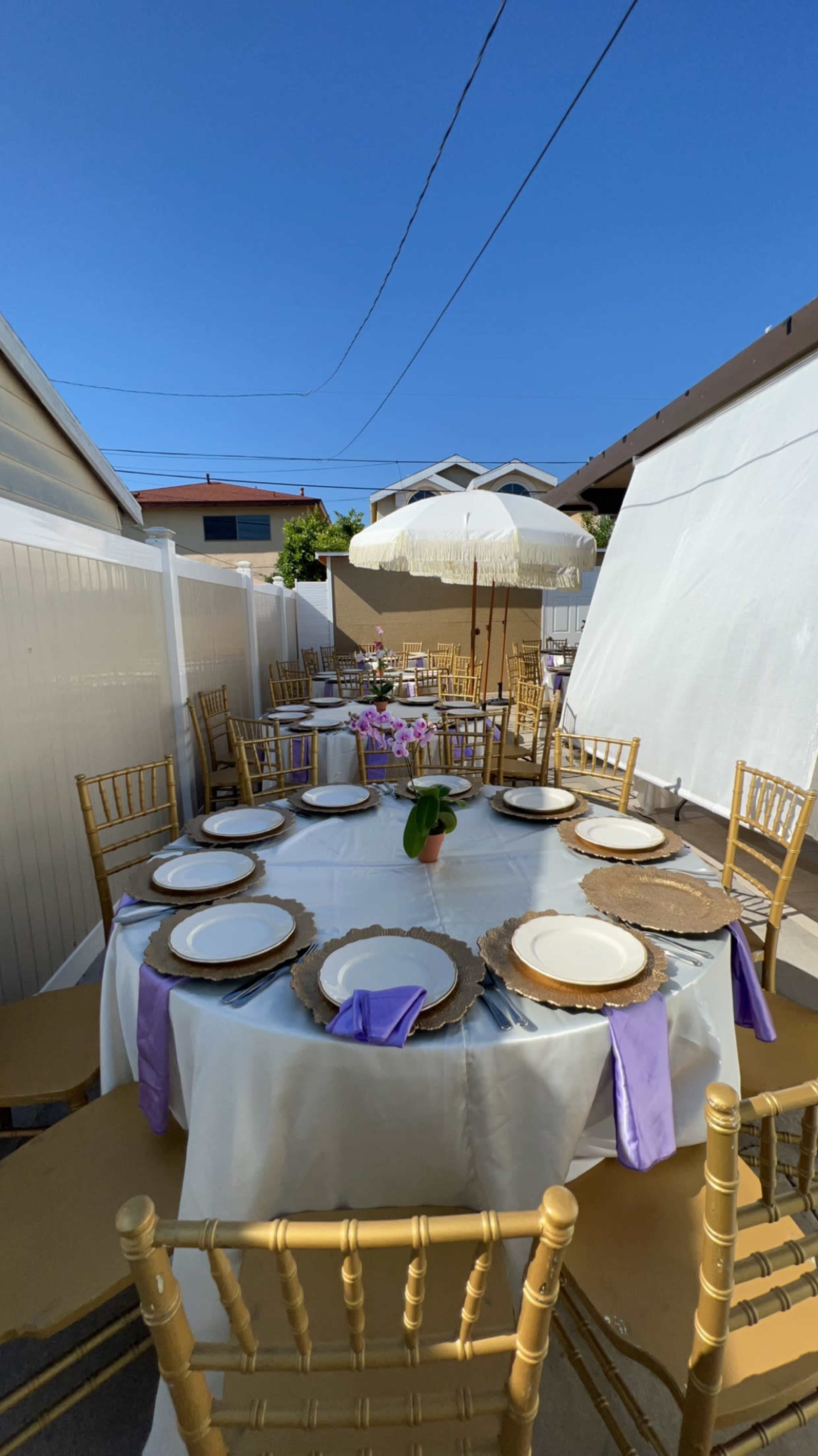 A backyard dining setup features several round tables adorned with white tablecloths, gold chairs, and purple napkins, under a large umbrella.