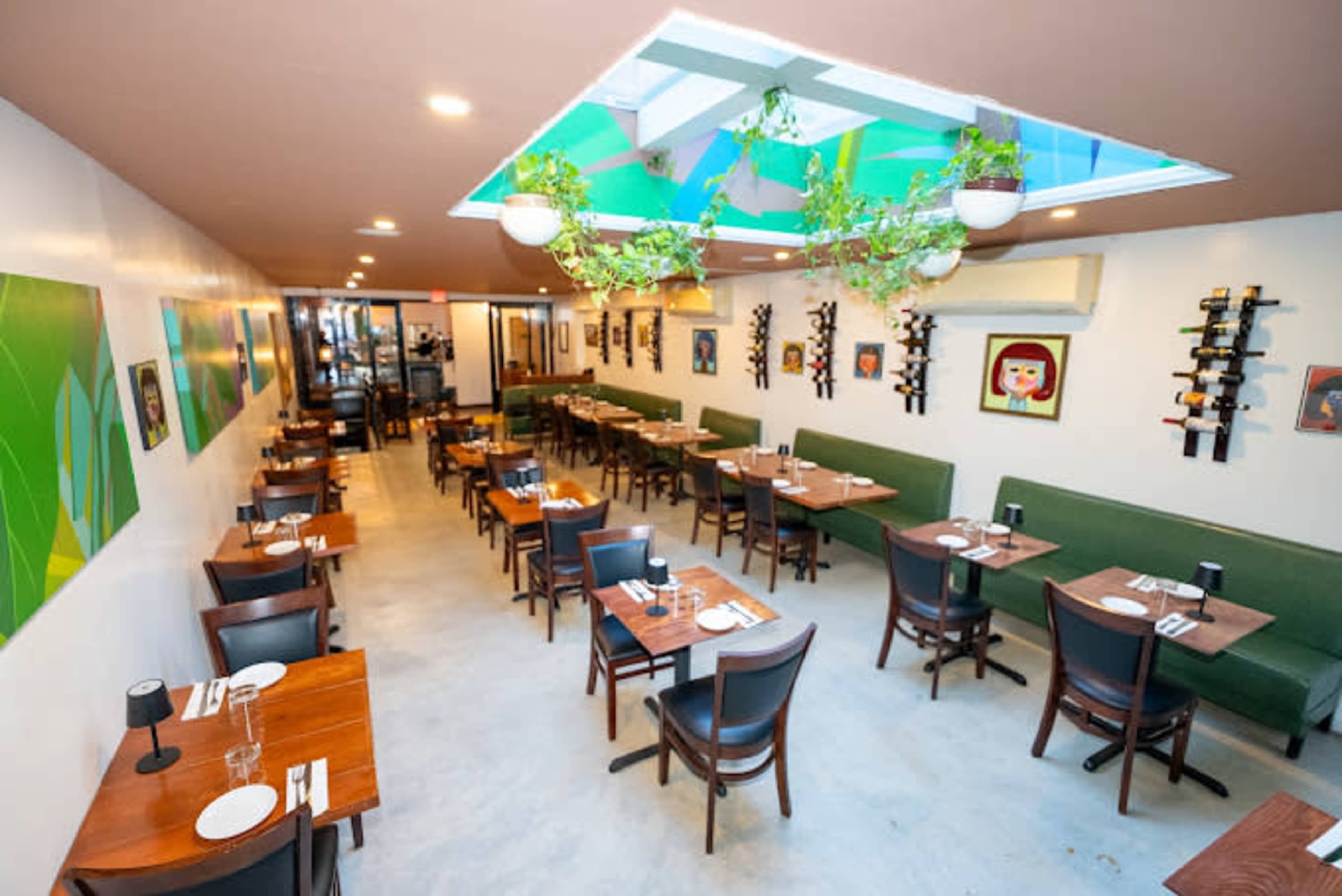 The image shows a modern restaurant interior with wooden tables and green seating, featuring artwork on the walls and plants hanging from the skylight.