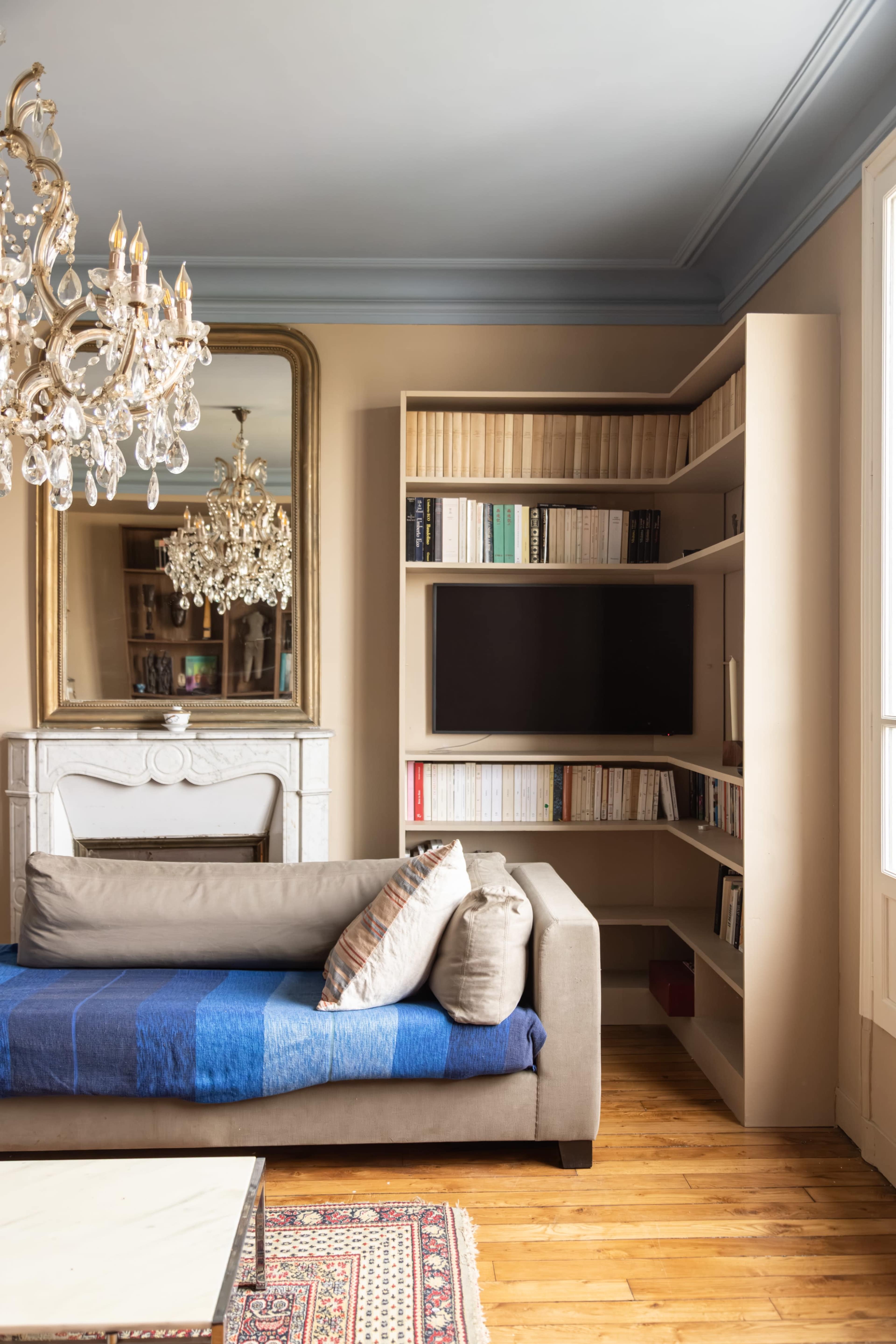 A modern living room features a beige sofa with colorful cushions, a shelving unit with books, and a mirror above a fireplace, illuminated by a chandelier.