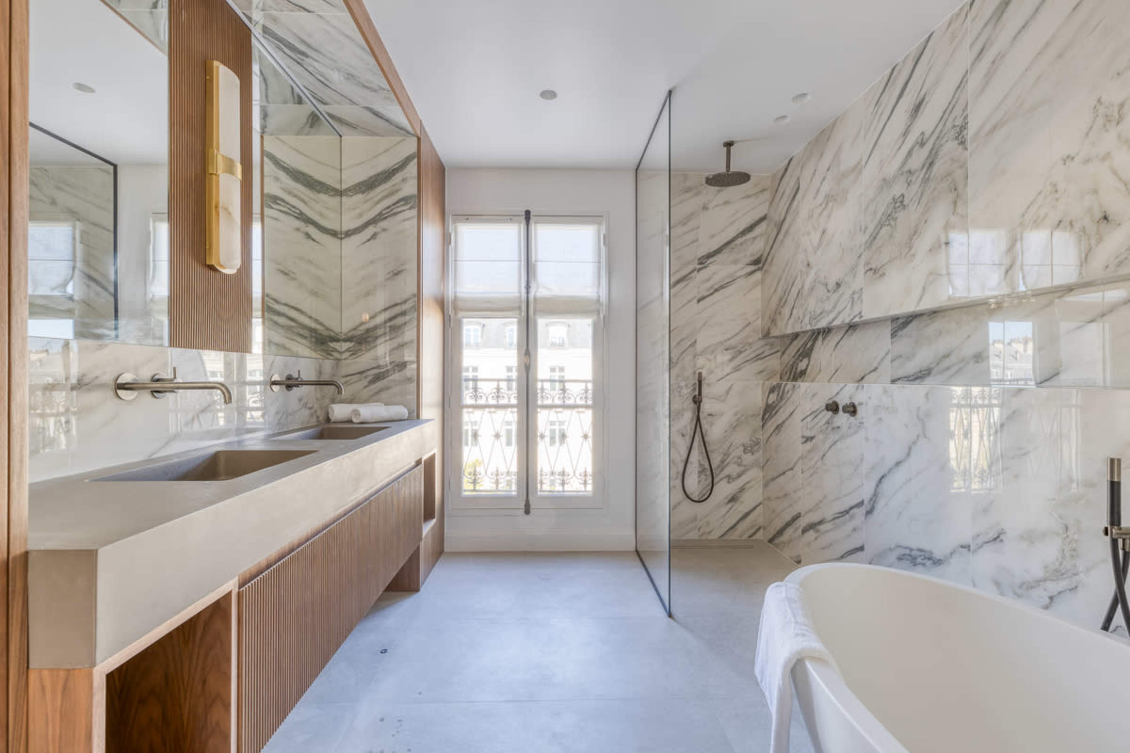The image shows a modern bathroom featuring a double sink vanity with wooden cabinetry, a freestanding bathtub, and a walk-in shower with marble walls.