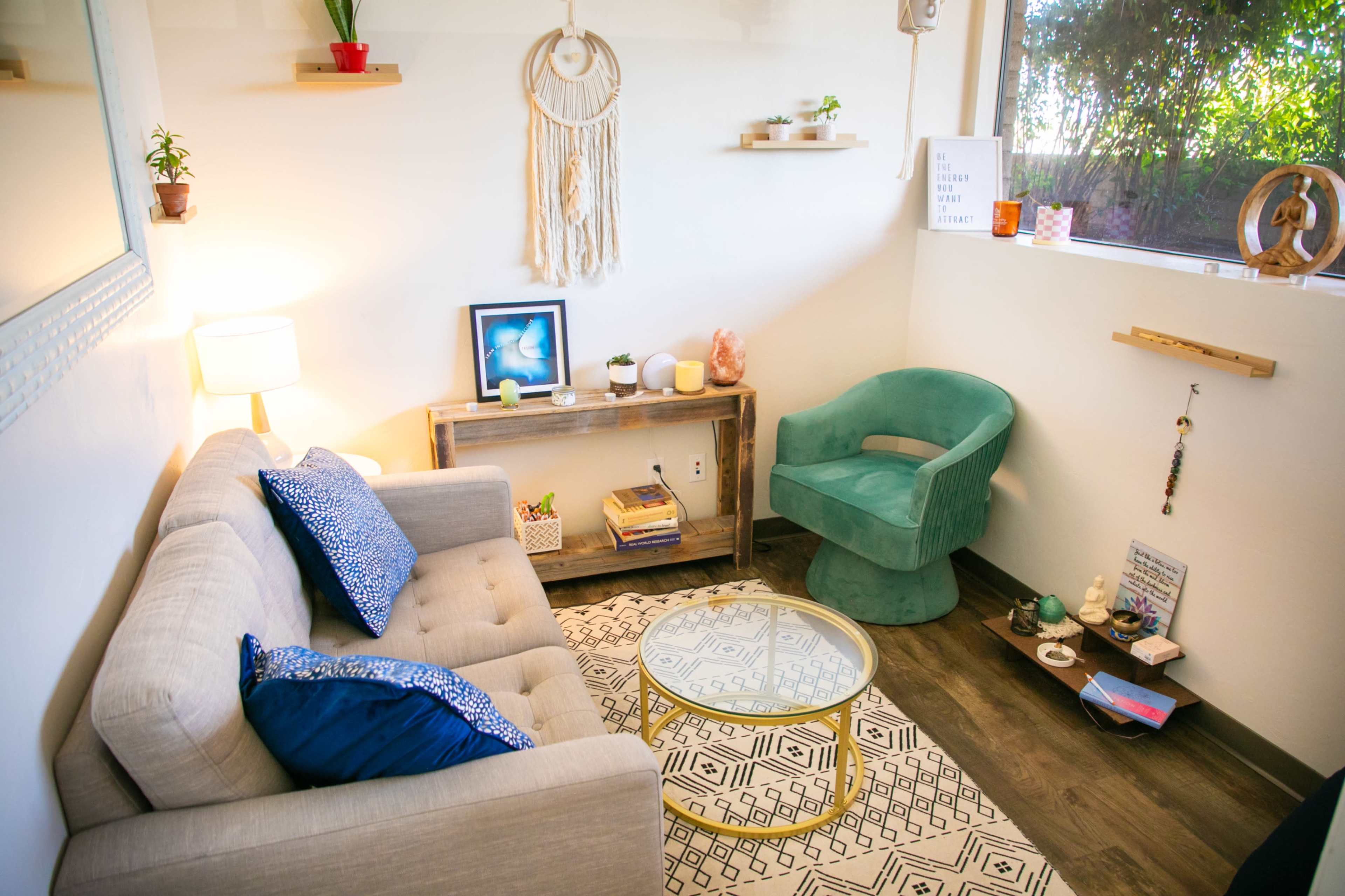 A cozy living room with a light-colored sofa, a glass coffee table, a wooden side table, and decorative plants on the shelves.