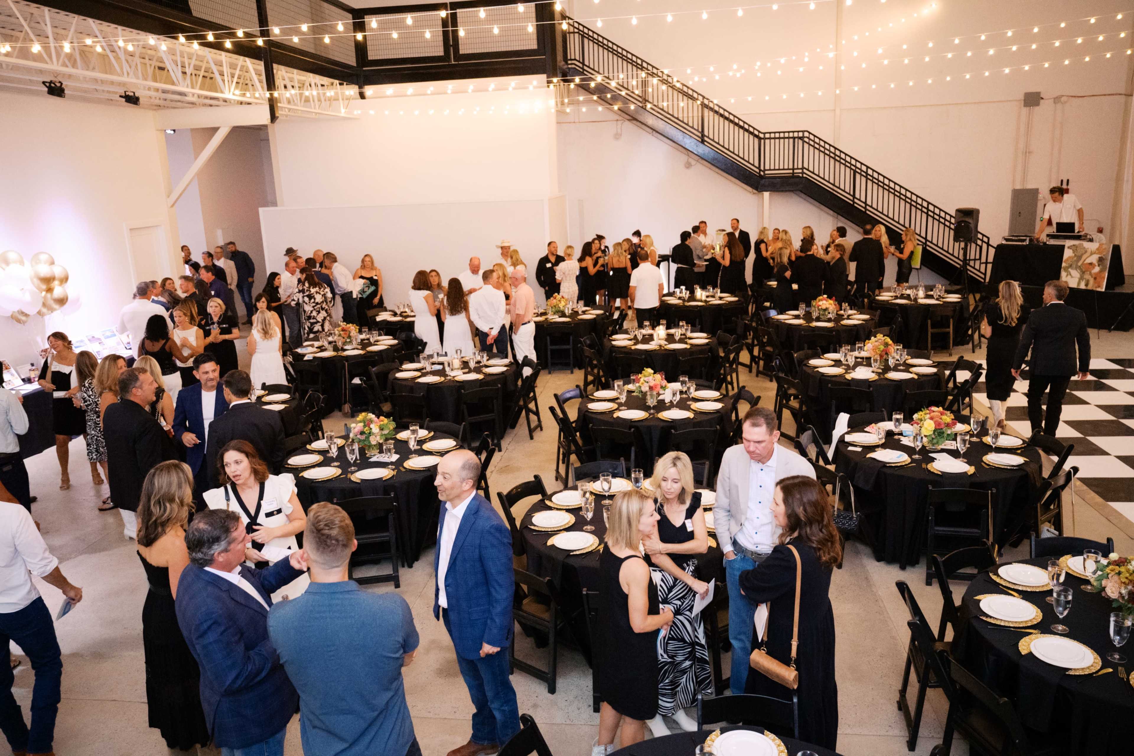 A large event is taking place in a spacious indoor venue with black tablecloths and string lights overhead, as guests mingle and socialize.