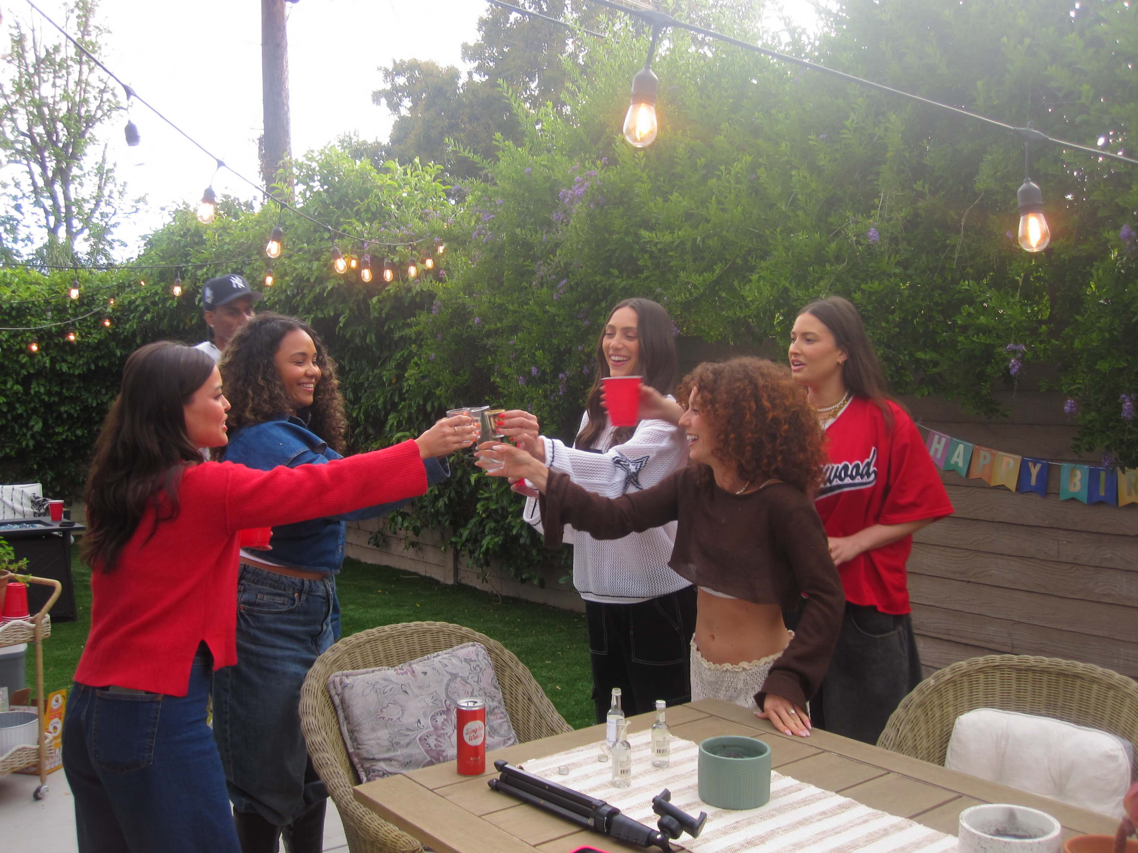 A group of five women and one man celebrate together in a backyard, raising their glasses for a toast under string lights.