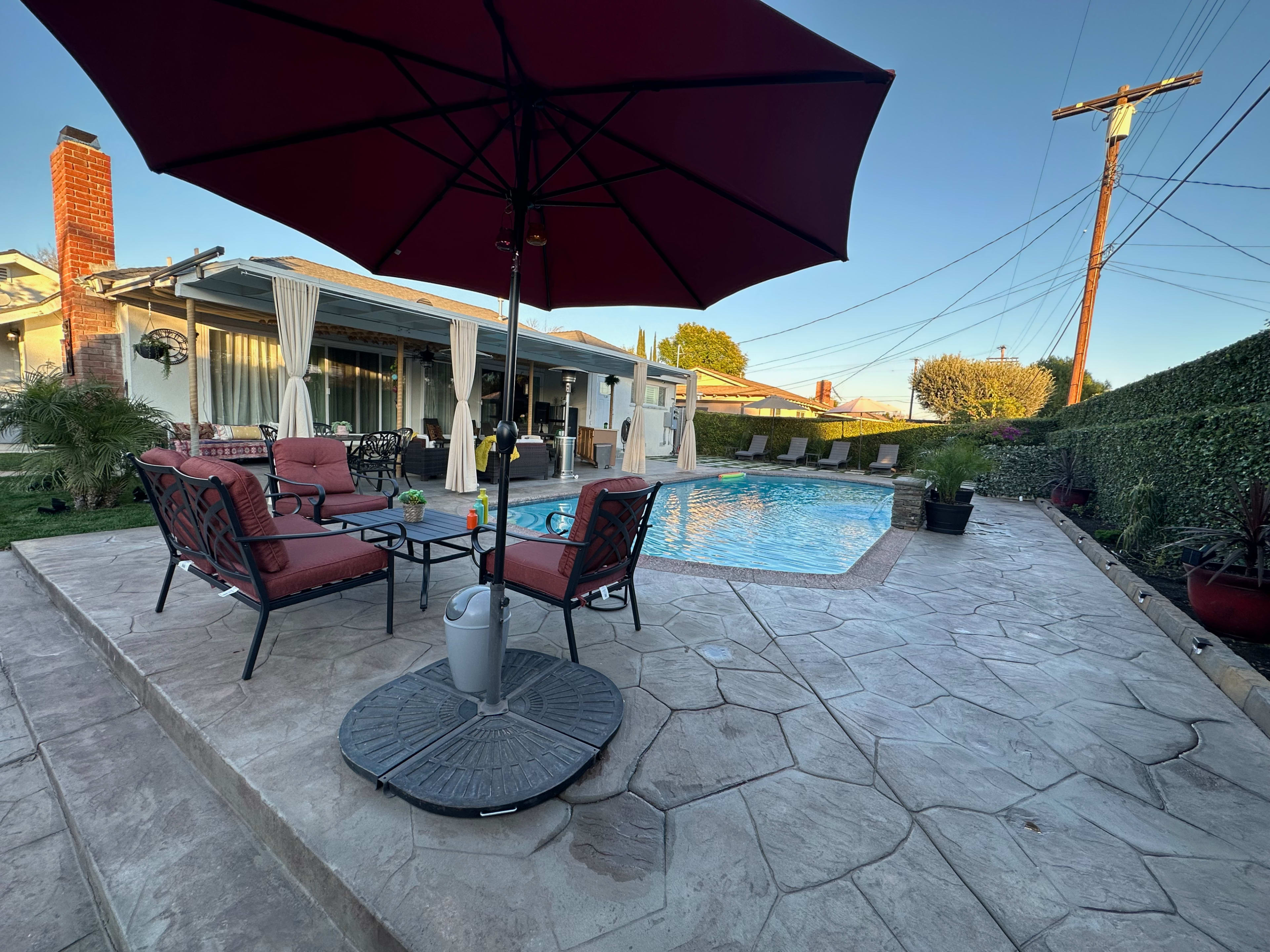 A swimming pool surrounded by patio seating and an umbrella, with a house and power lines in the background.