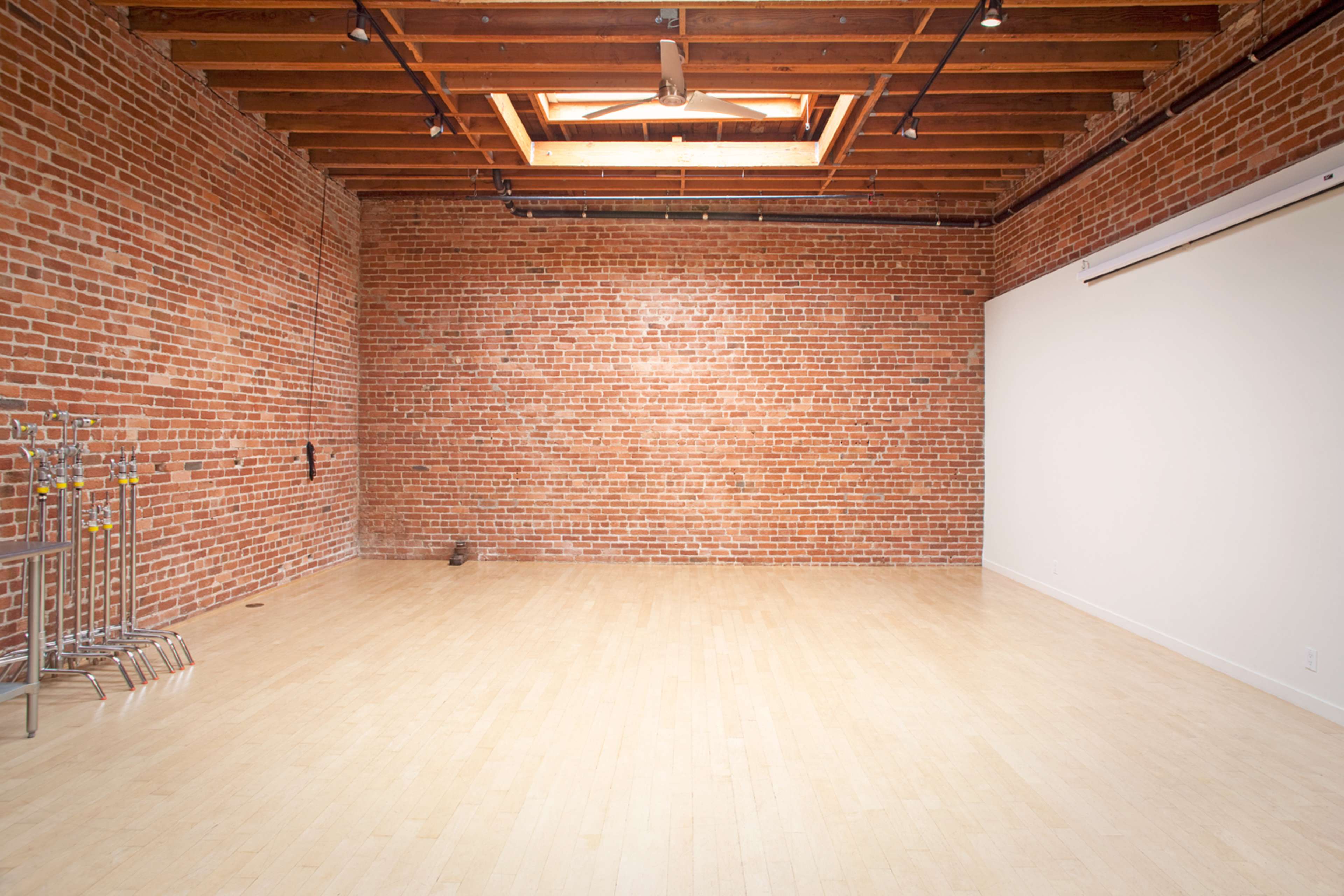 A spacious room with exposed brick walls, wooden beams, and a light wooden floor, featuring a skylight at the ceiling.