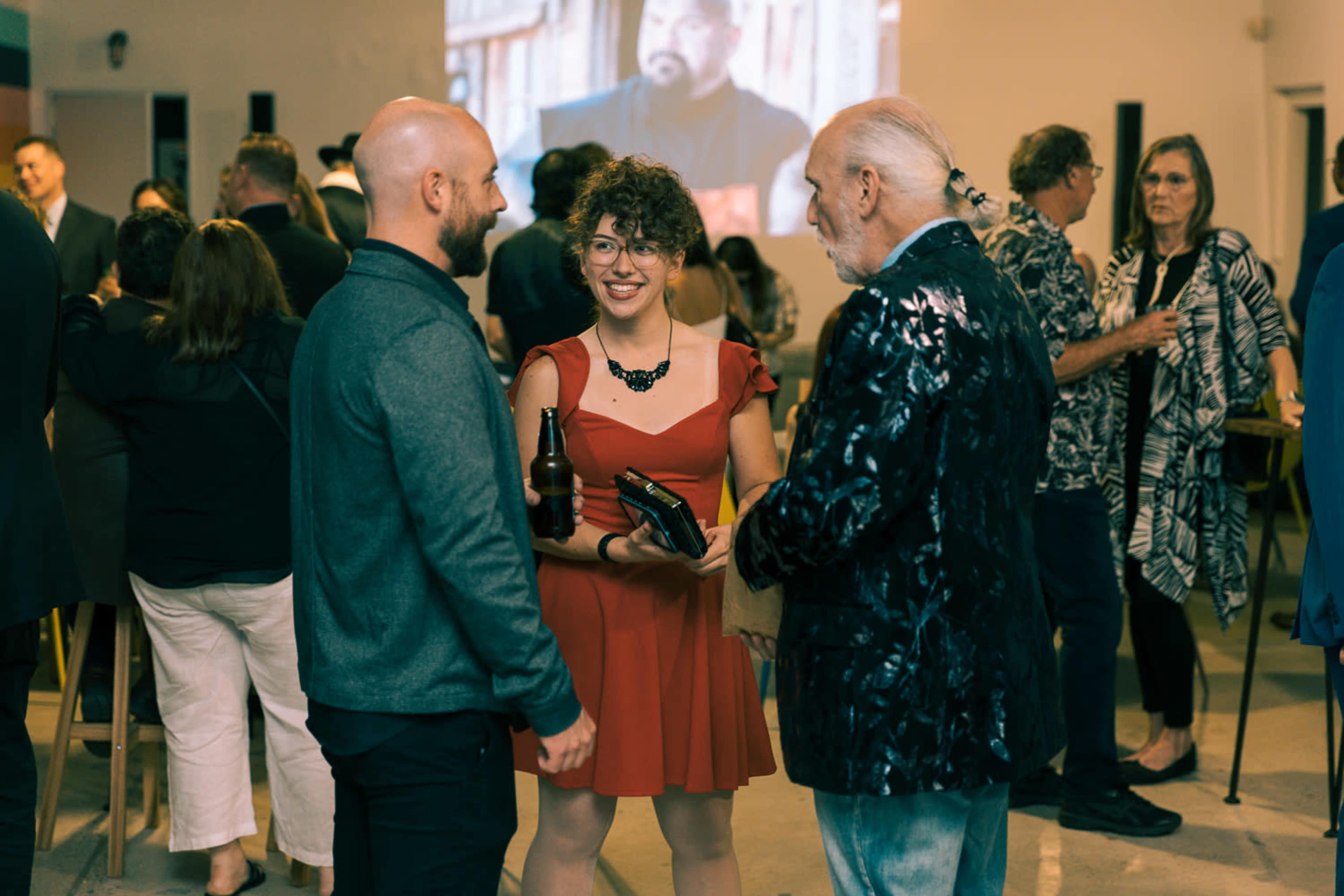 A group of people engages in conversation at a social event, with a woman in a red dress holding a drink while standing between two men.