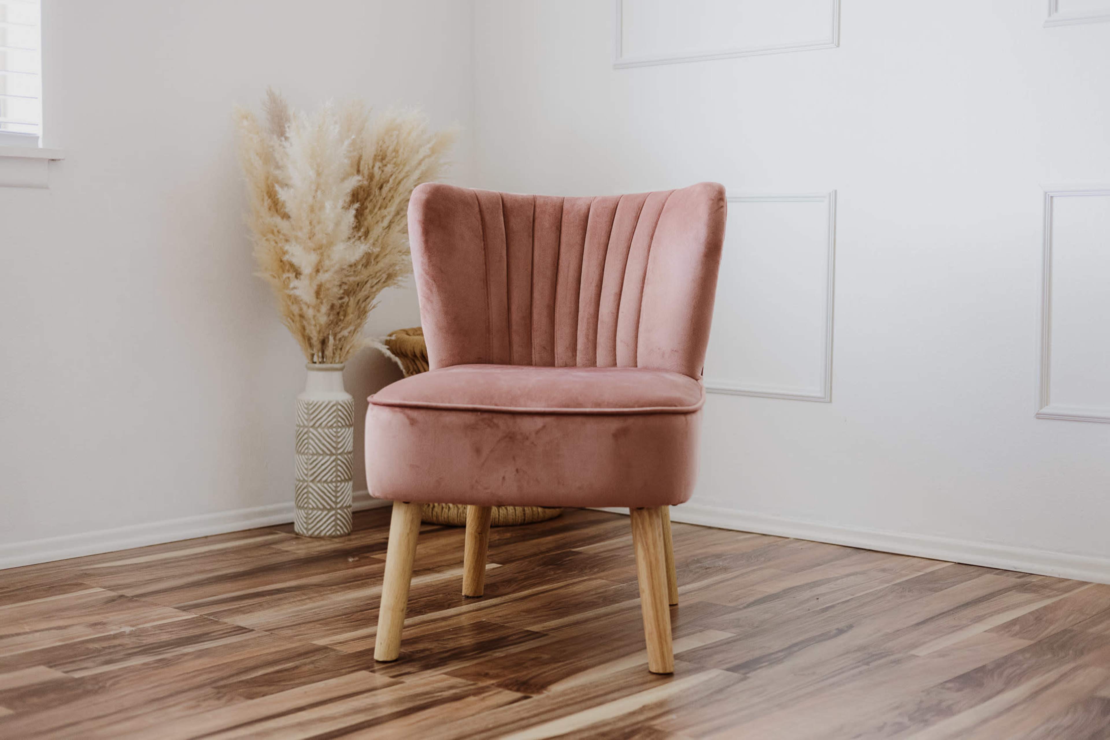A pink upholstered chair with vertical pleats sits next to a decorative vase of dried pampas grass on a wooden floor.