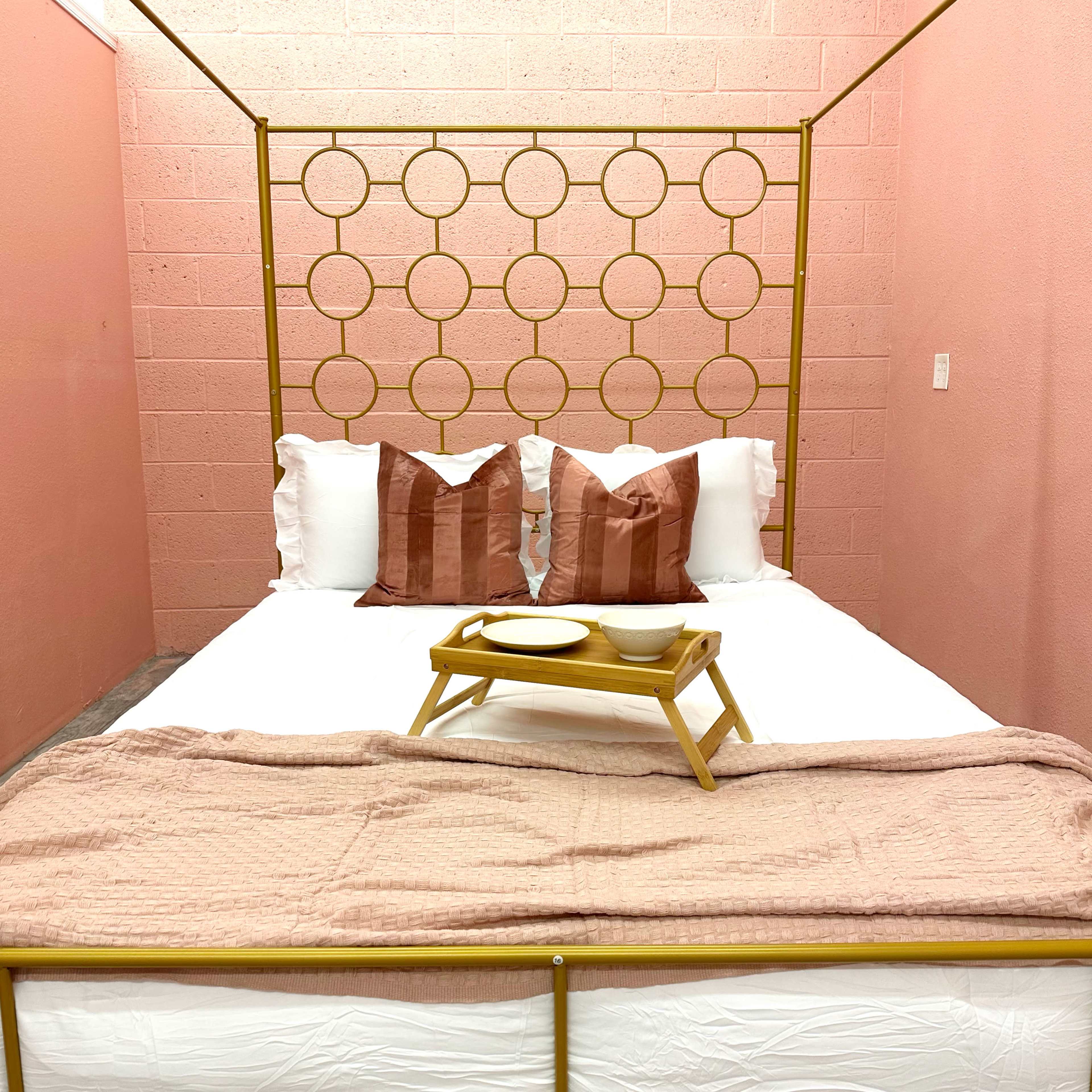 A neatly arranged bed with a gold metal frame and pink accents is set against a pink wall, featuring decorative pillows and a tray with dishes on top.