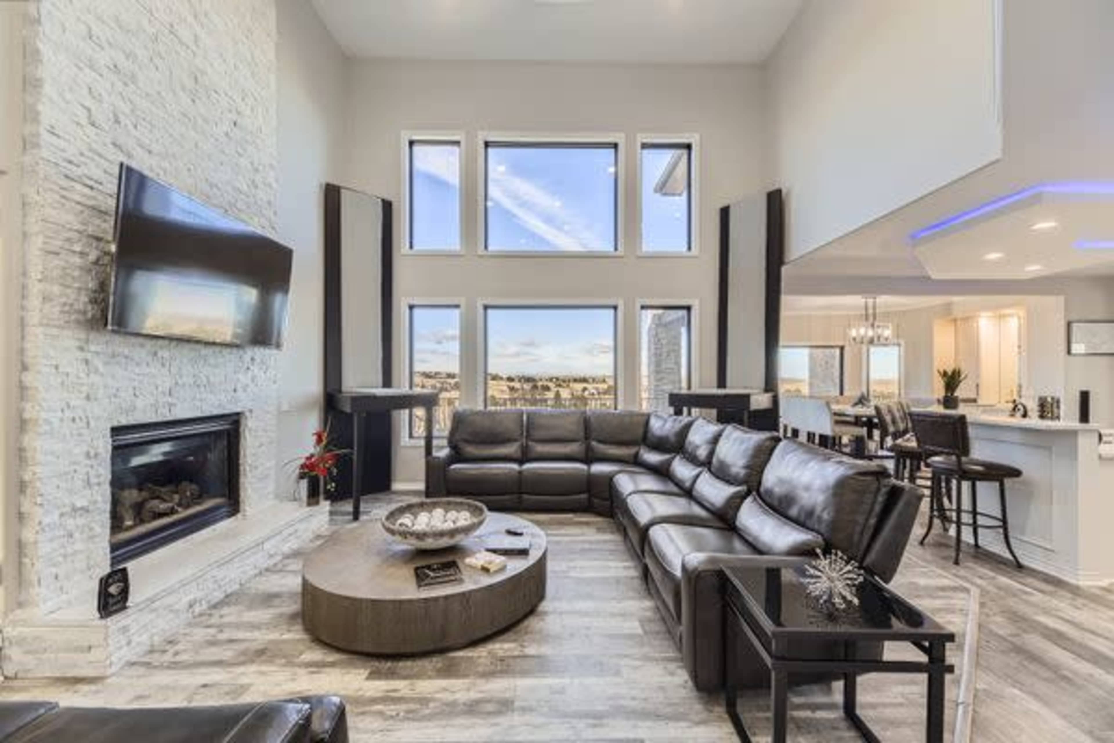 A modern living room features a large sectional sofa, a circular coffee table, a stone fireplace, and expansive windows overlooking a scenic view.