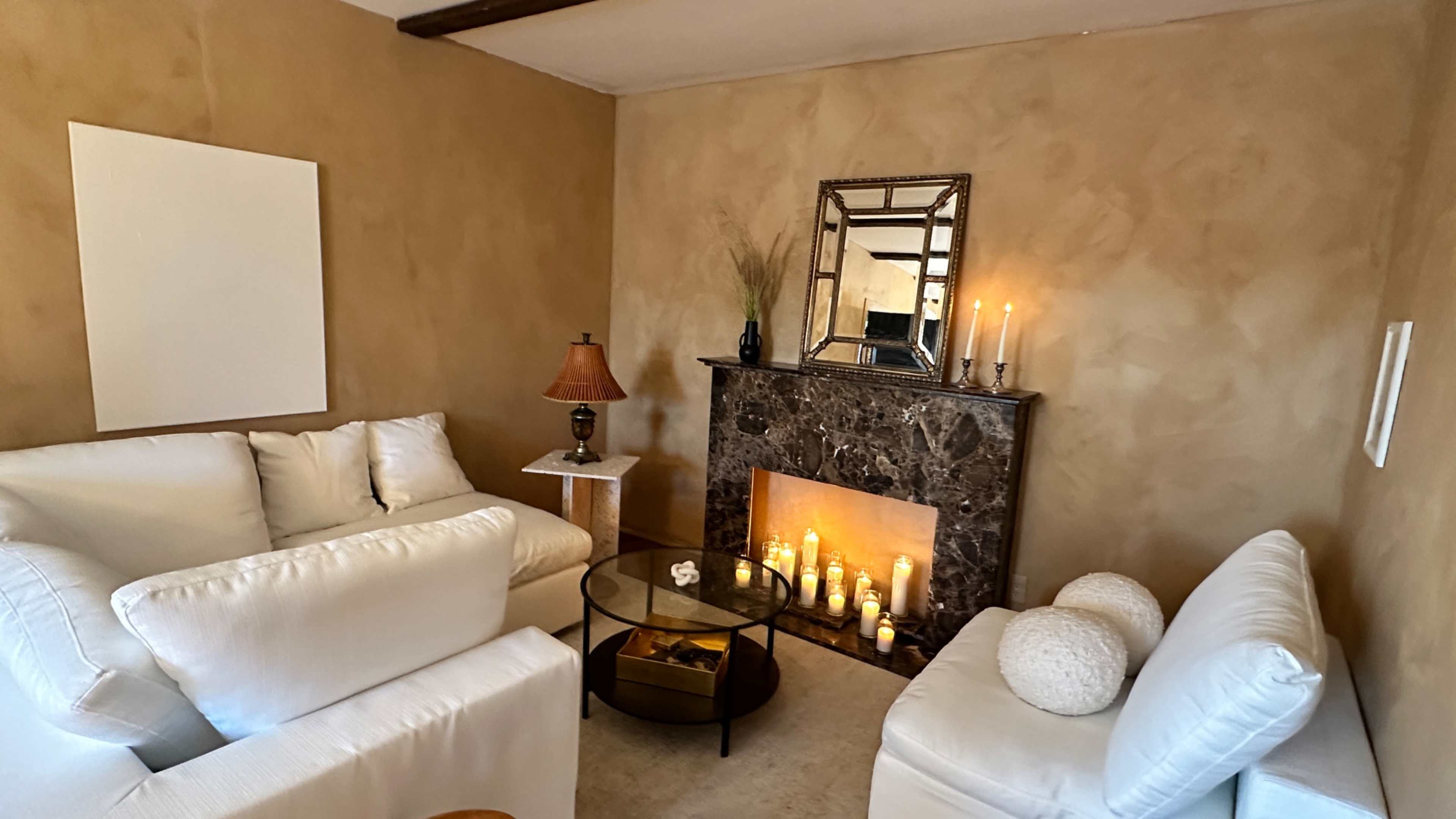 A cozy living room with two white sofas facing a dark marble fireplace adorned with candles and a mirror above it.