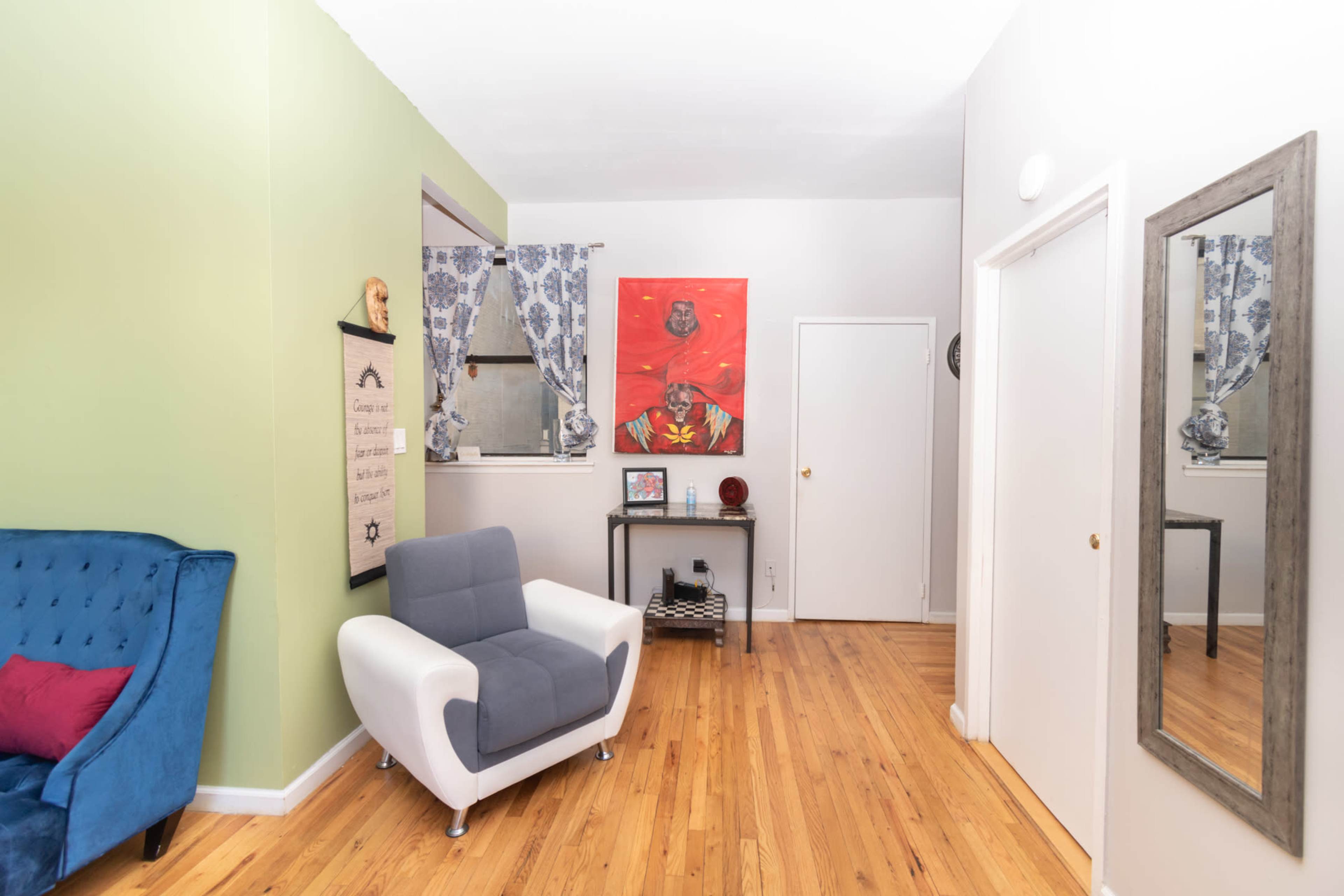 A small living area featuring a blue couch and a gray accent chair, with a colorful artwork on the wall and wooden flooring.