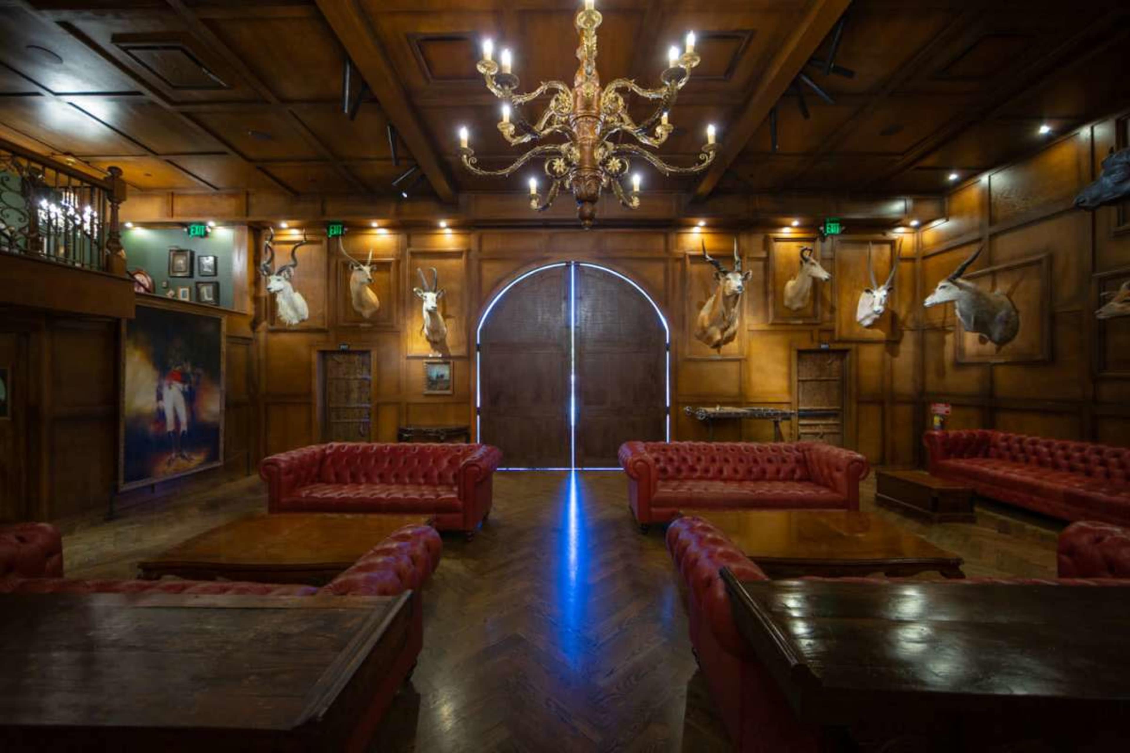 The room features wooden paneling, leather sofas arranged around wooden tables, and animal heads mounted on the walls, illuminated by a large chandelier.