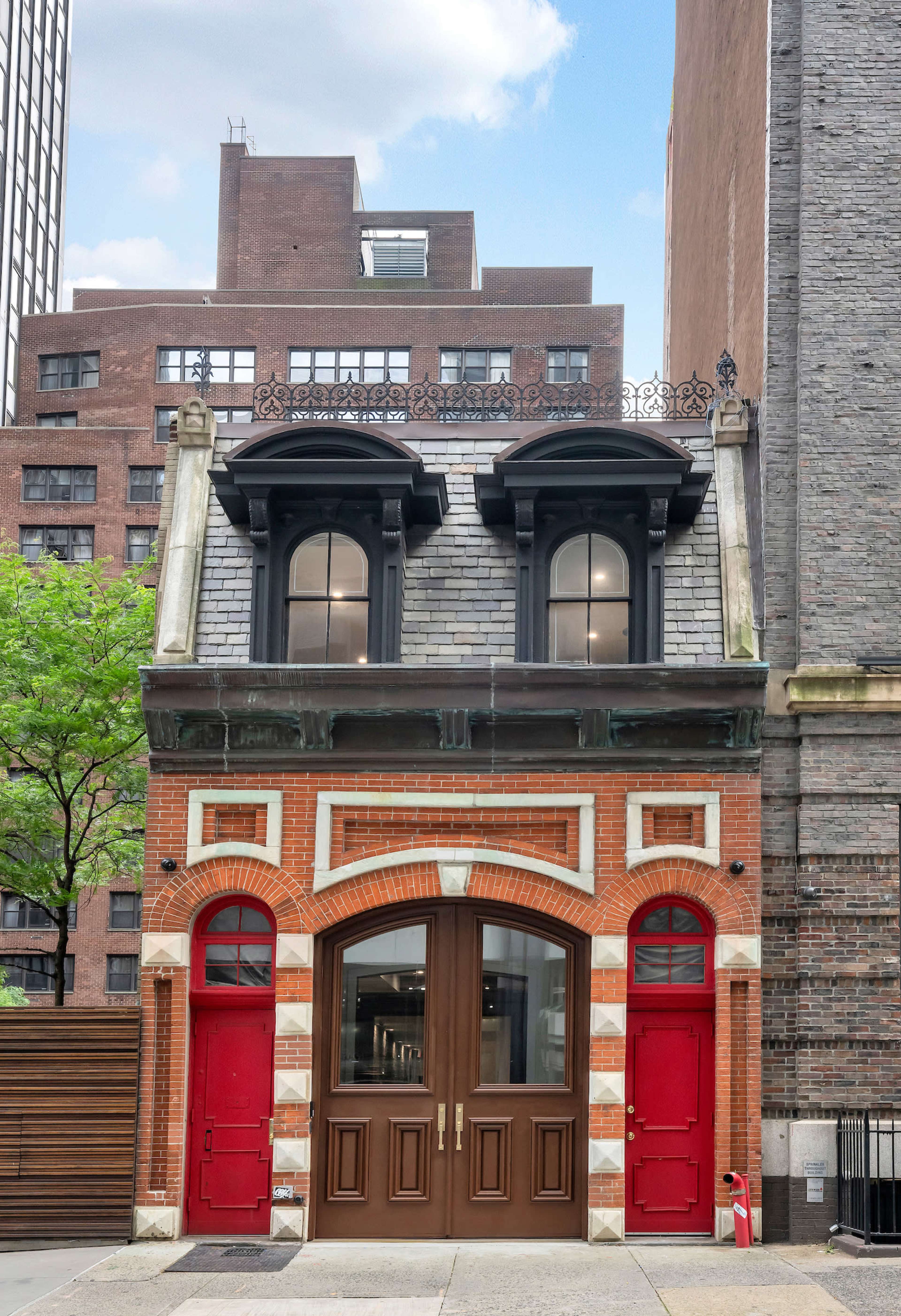 A historic building with a red brick facade, two arched doors, and a decorative roof, situated between modern structures in an urban setting.