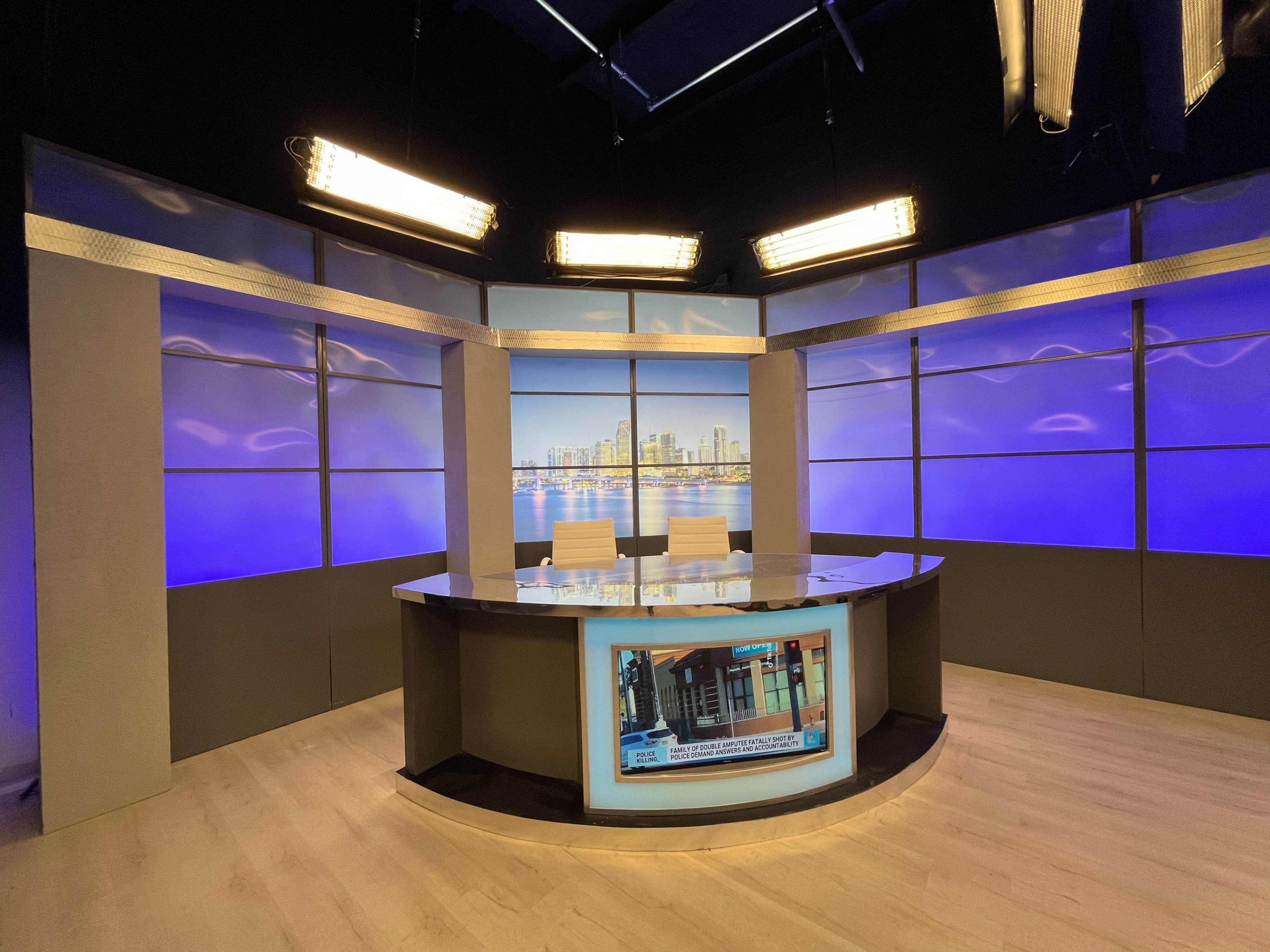 A television news studio features a modern desk setup with two chairs and large screens displaying a city skyline backdrop.