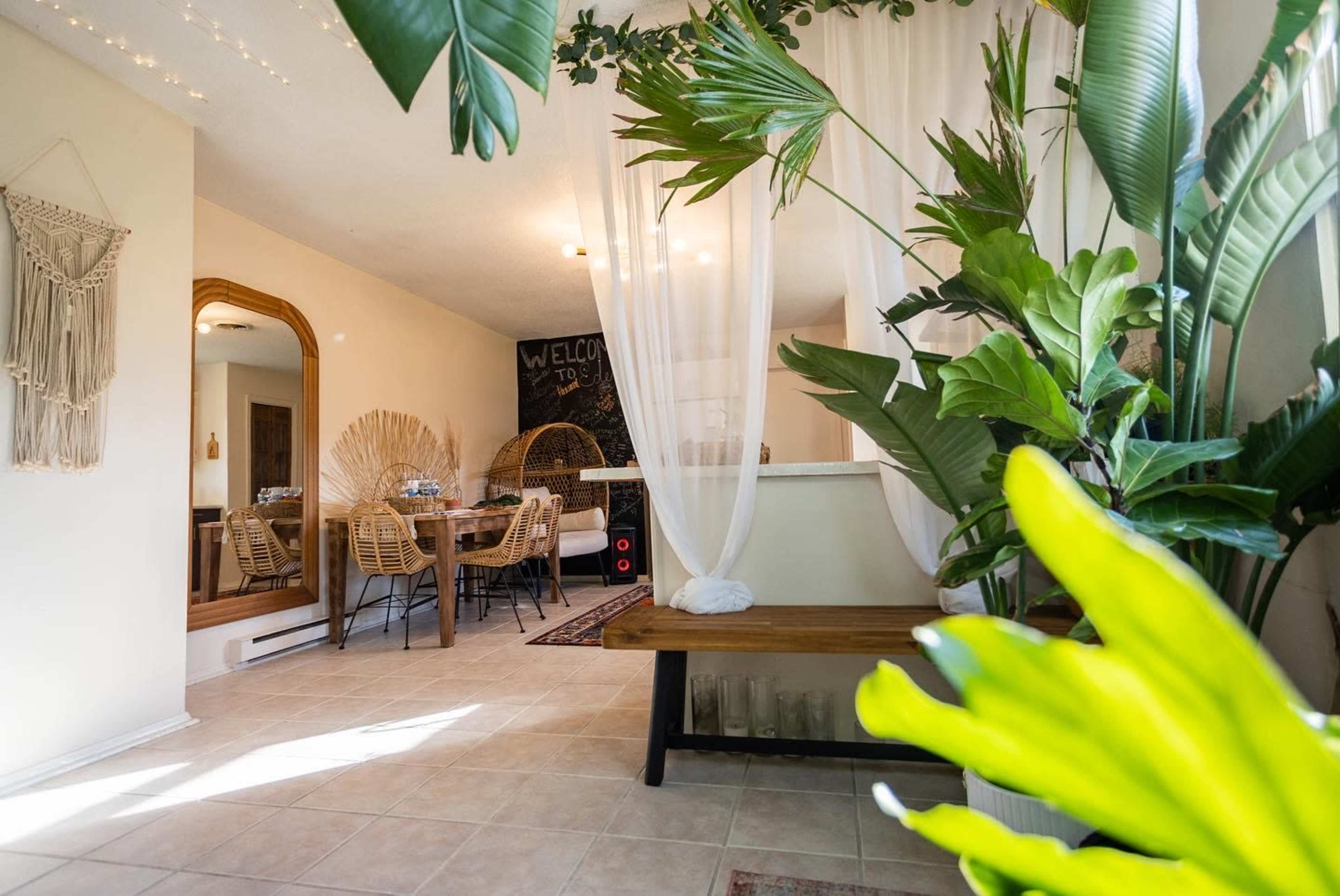 A cozy interior space with a beige tiled floor, featuring a bench with plants, a dining area with chairs, and a welcoming decor that includes hanging drapes and greenery.
