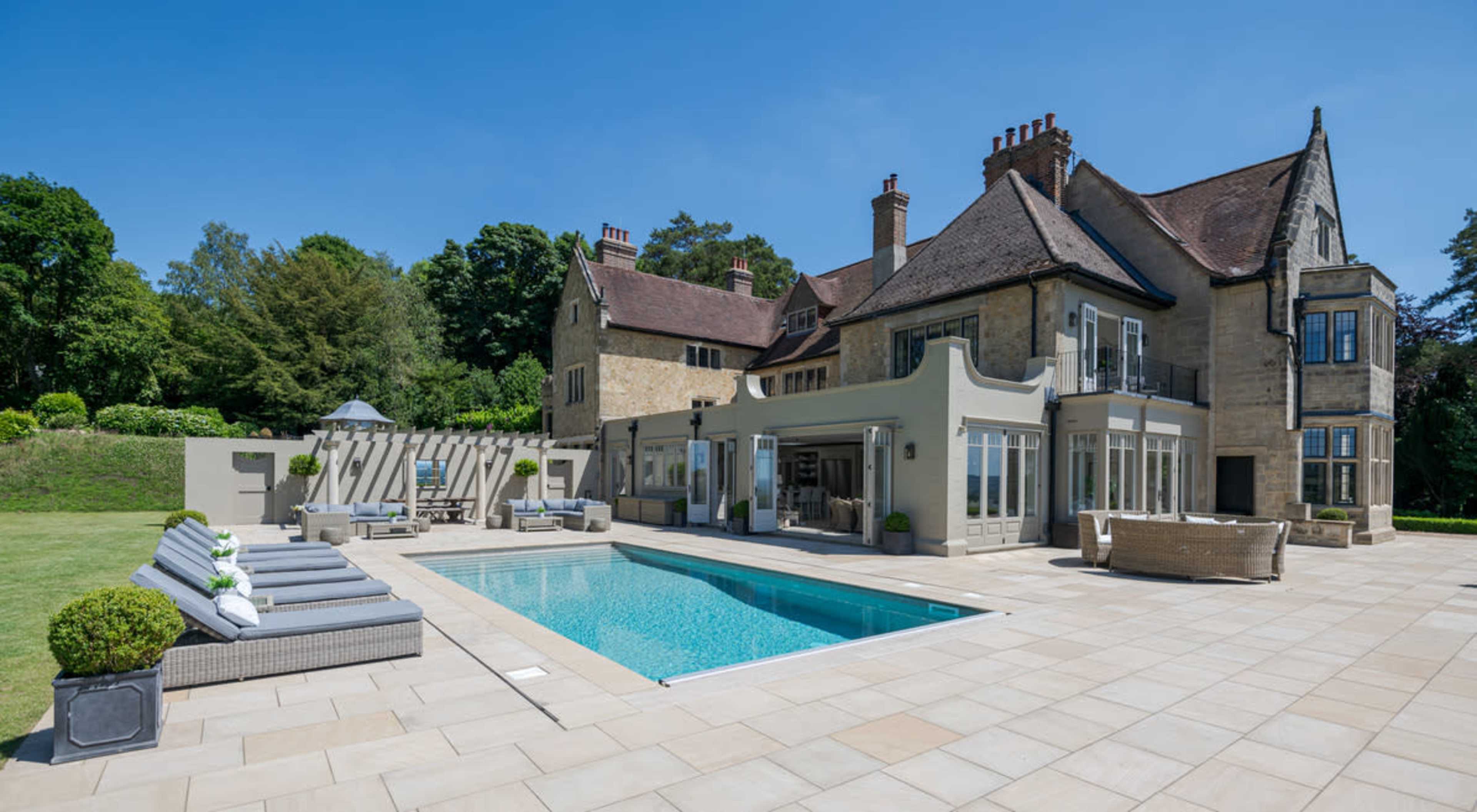 A luxurious stone house with a swimming pool and outdoor seating, surrounded by well-manicured lawns and trees.