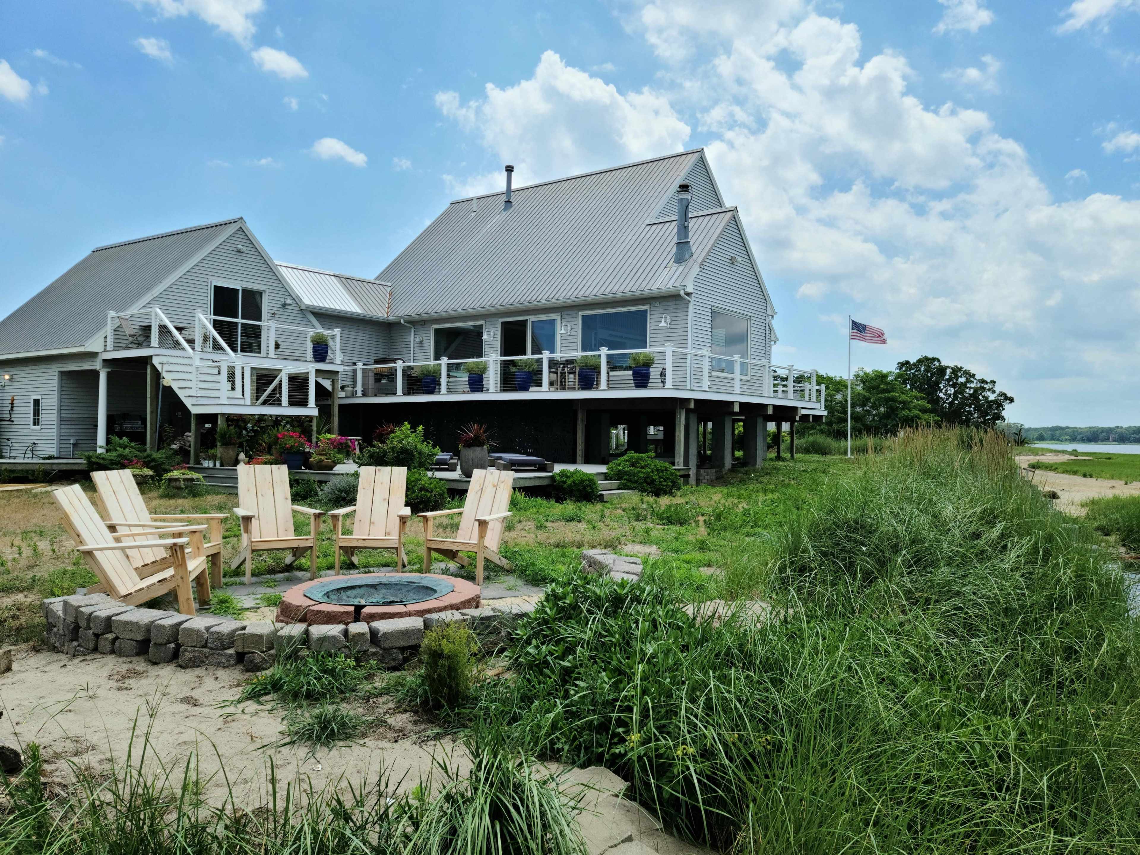 A modern house with a deck overlooks a fire pit surrounded by Adirondack chairs, set against a backdrop of grass and water.