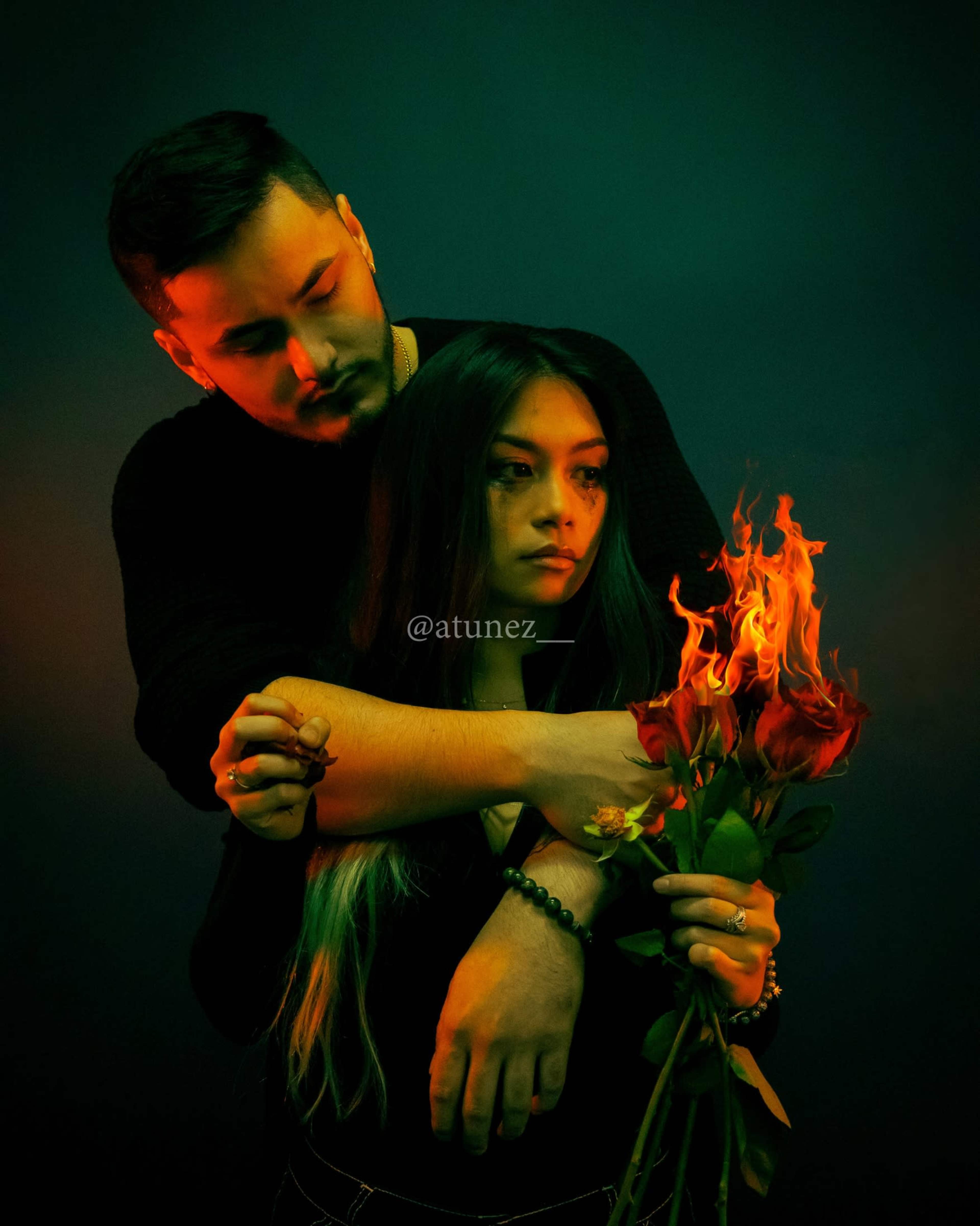A man holds a woman from behind as she stands with a bouquet of roses, one of which is engulfed in flames.