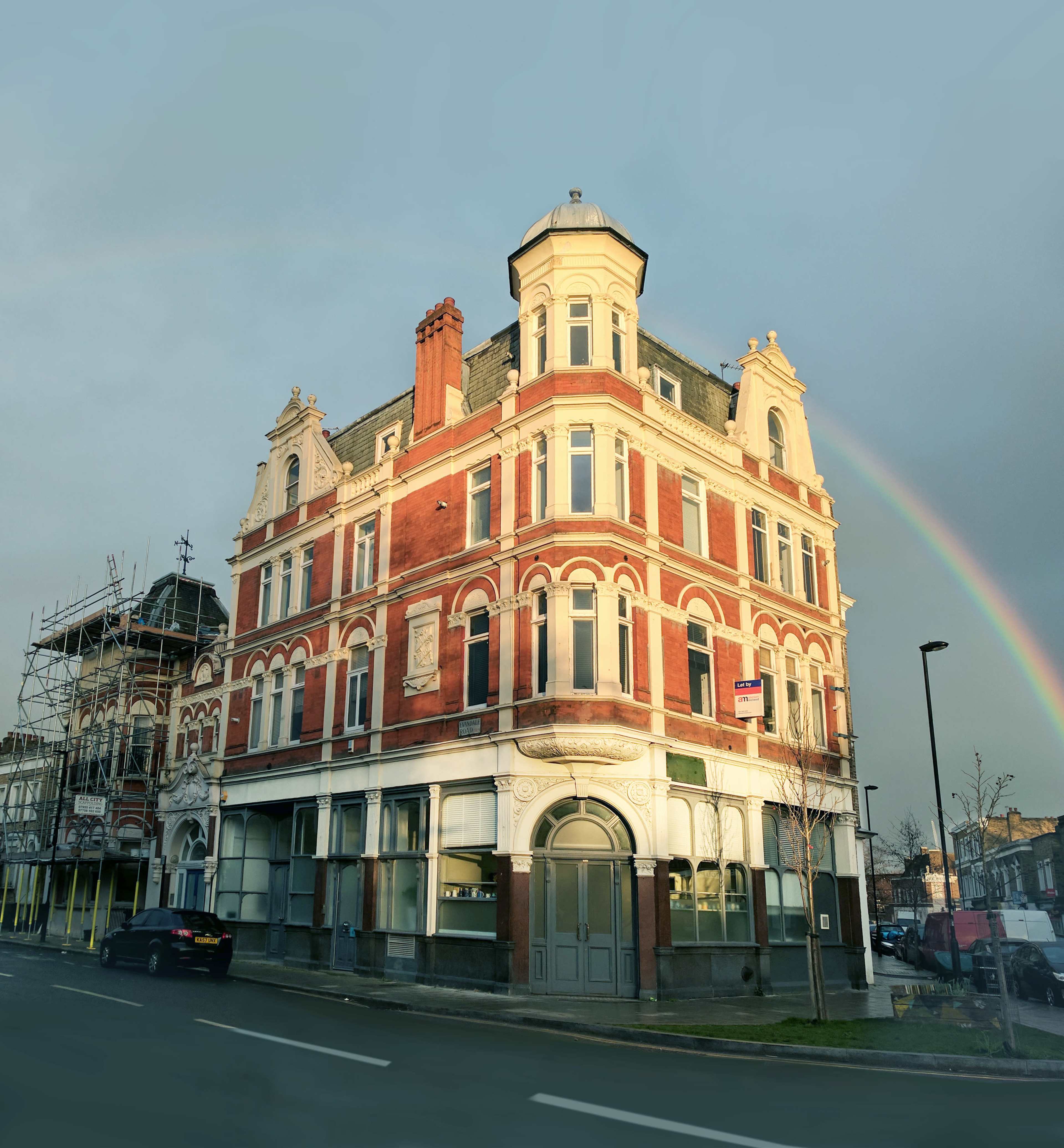 A three-story brick building with ornate architecture stands at a street corner, featuring scaffolding on one side and a rainbow in the sky above.