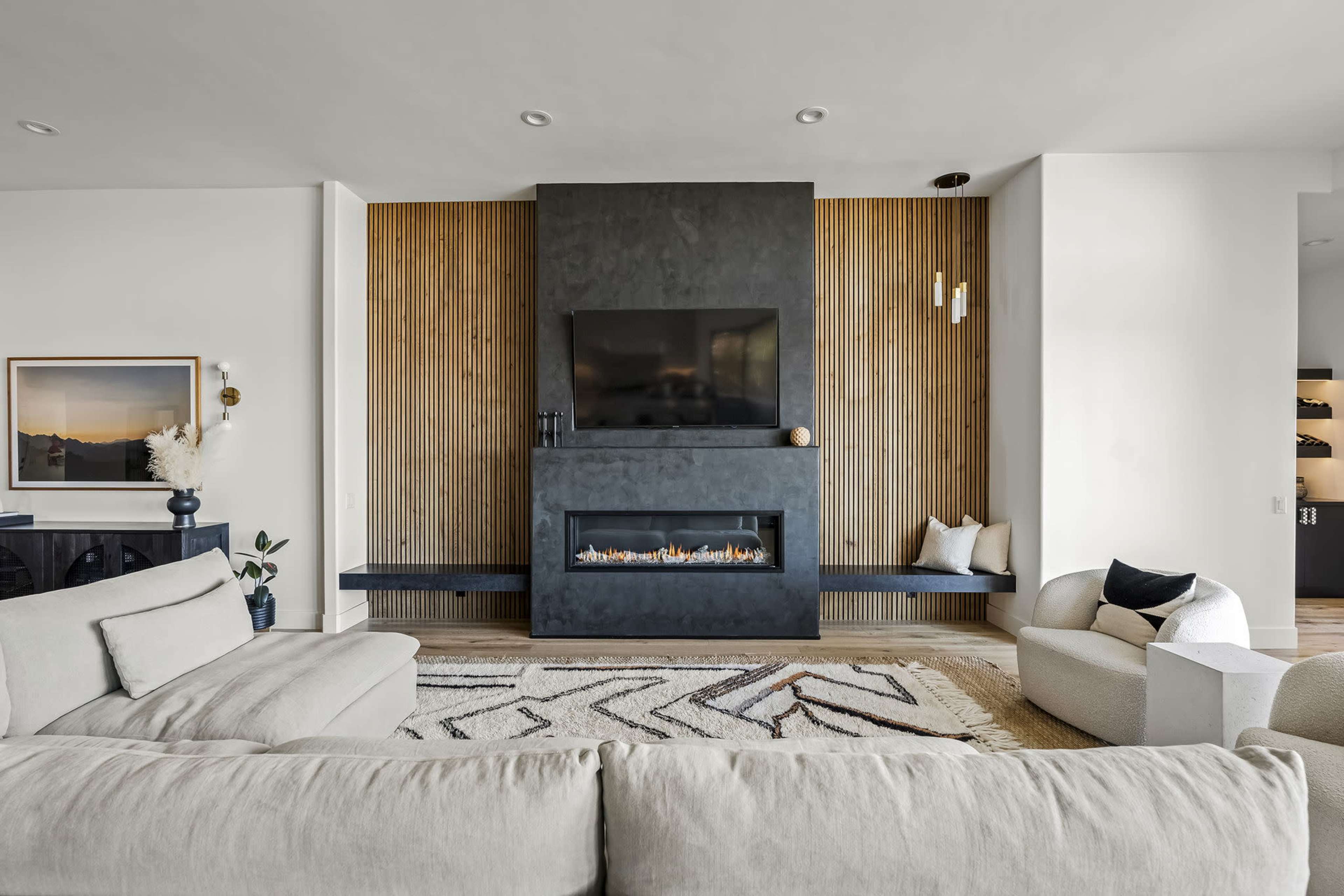 A modern living room with a flat-screen TV mounted above a sleek fireplace, flanked by a wooden wall and decorated with minimal furniture.
