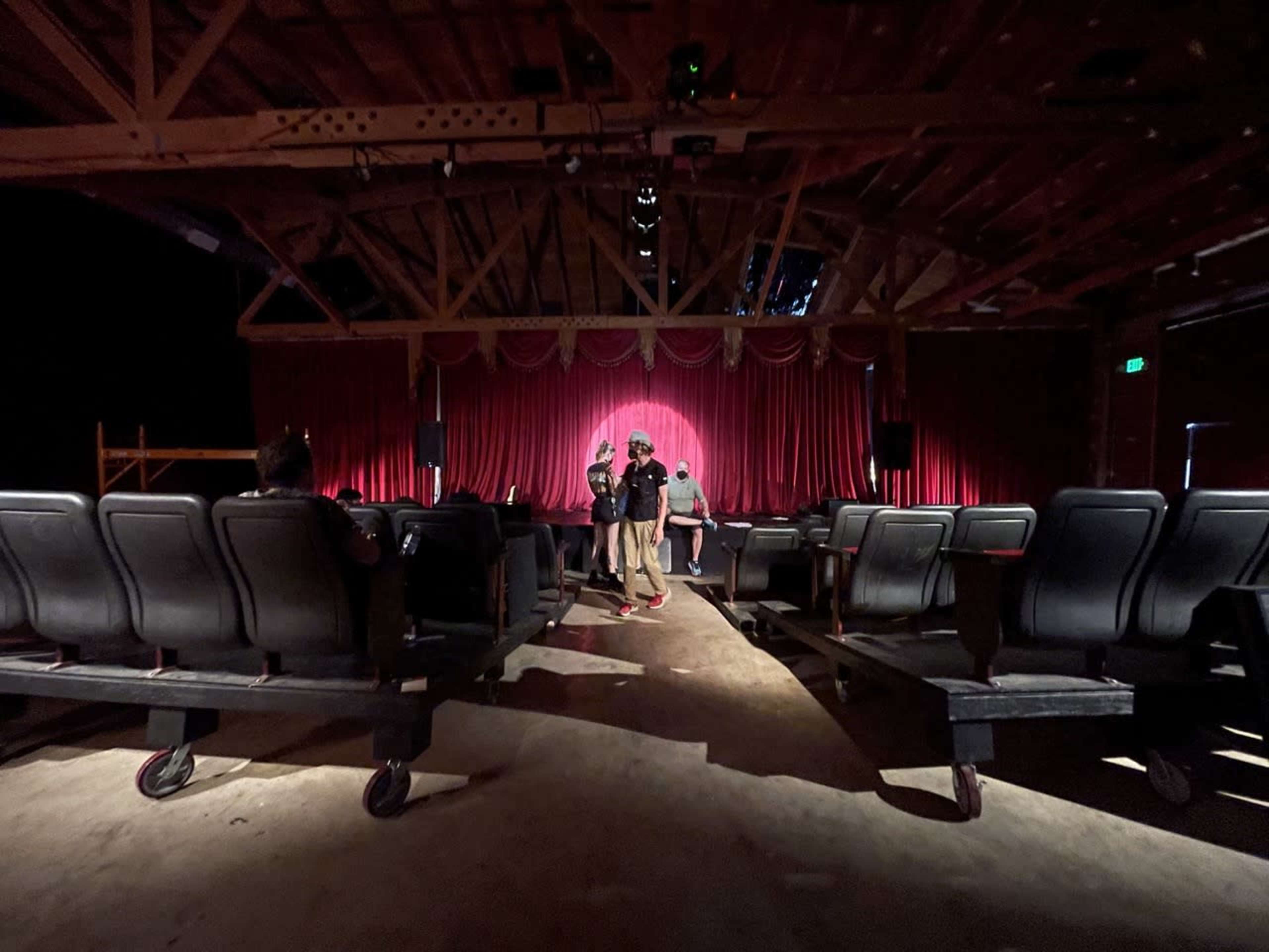 A small theater with rows of empty chairs faces a red curtain, where a few people are gathered on stage.