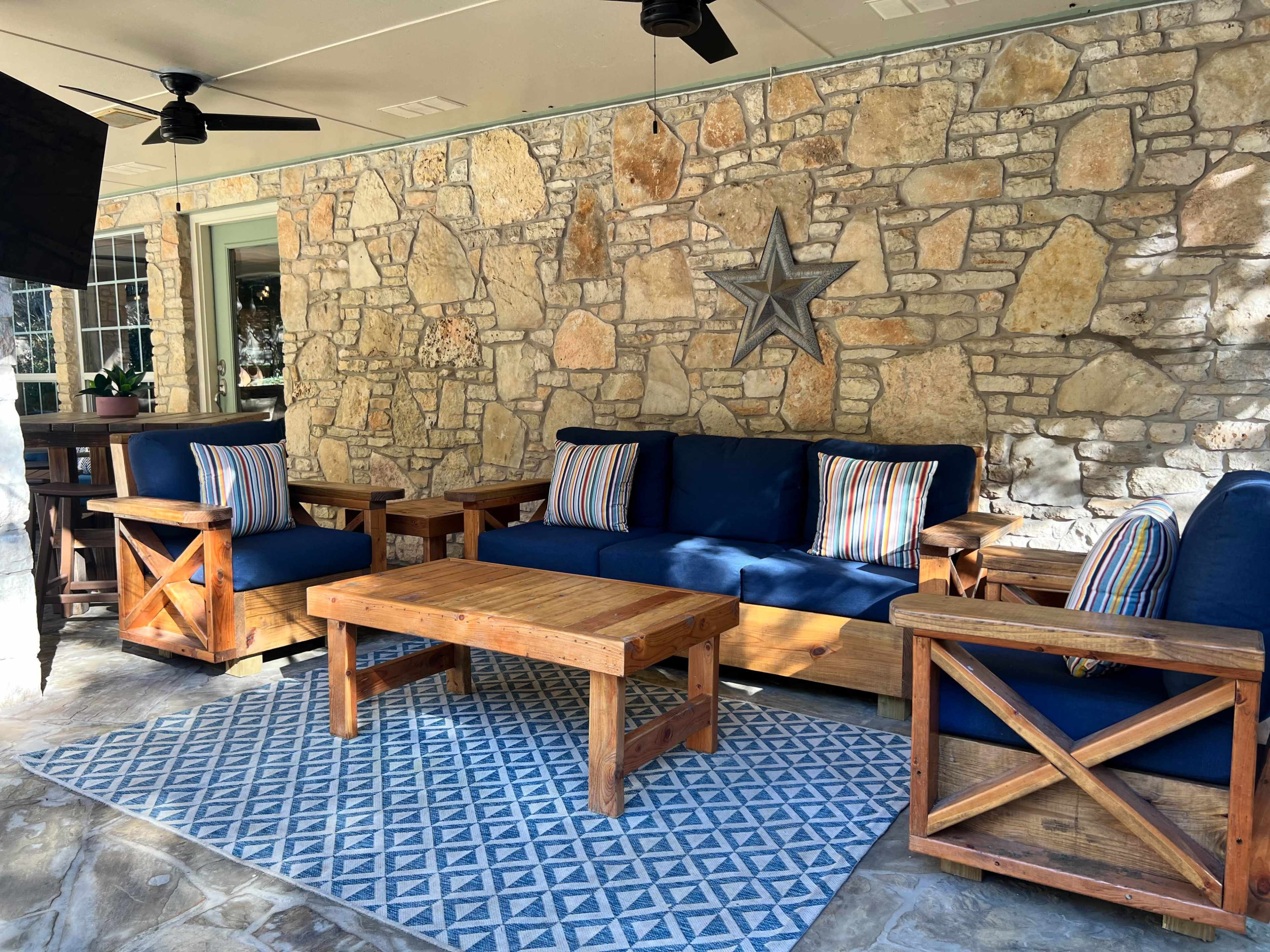 The image shows a cozy outdoor seating area with a stone wall backdrop, featuring a wooden sofa with blue cushions, two matching chairs, and a wooden coffee table on a patterned rug.
