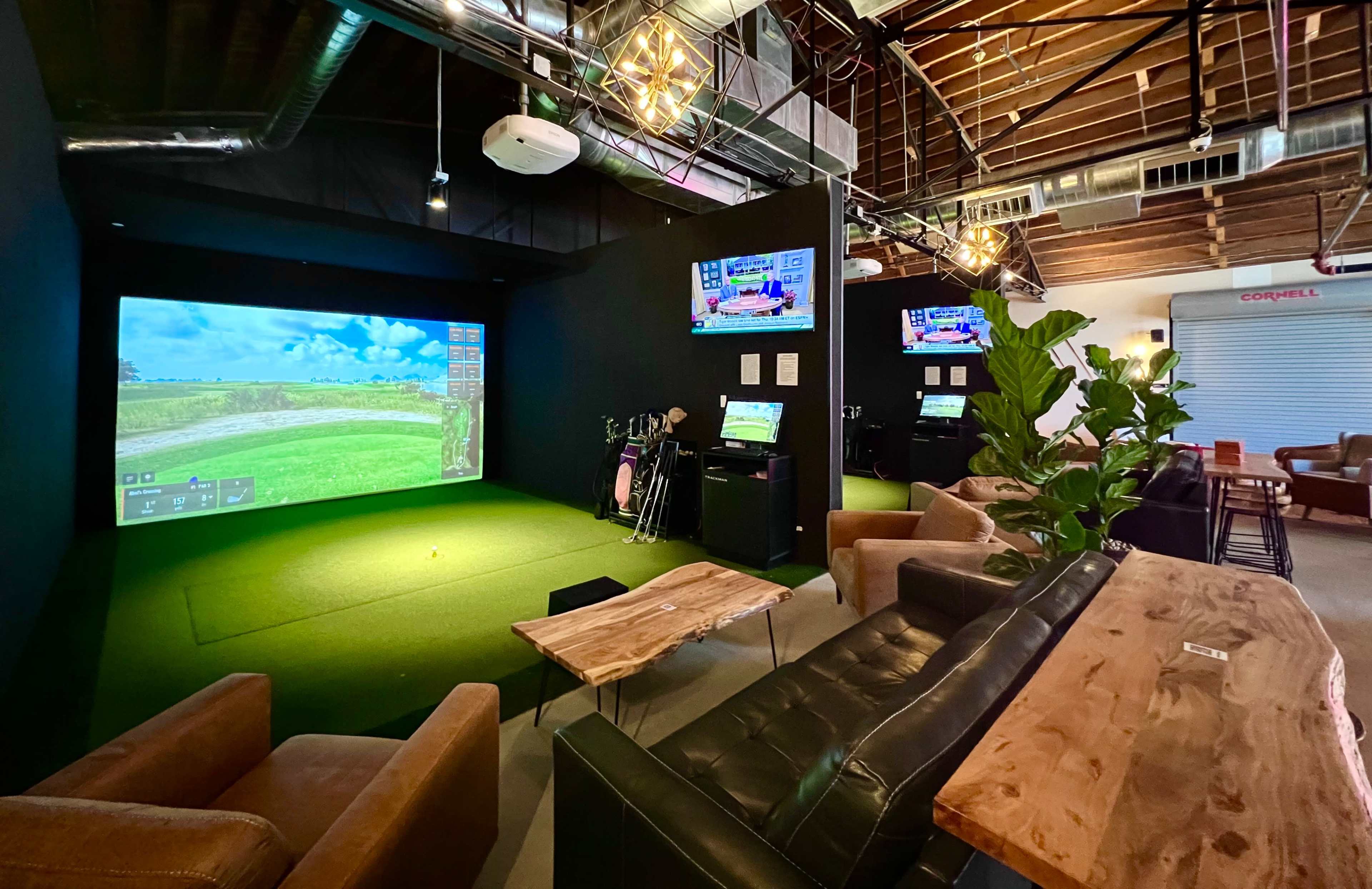An indoor golf simulation area with a large screen displaying a golf course, surrounded by seating and golf equipment.