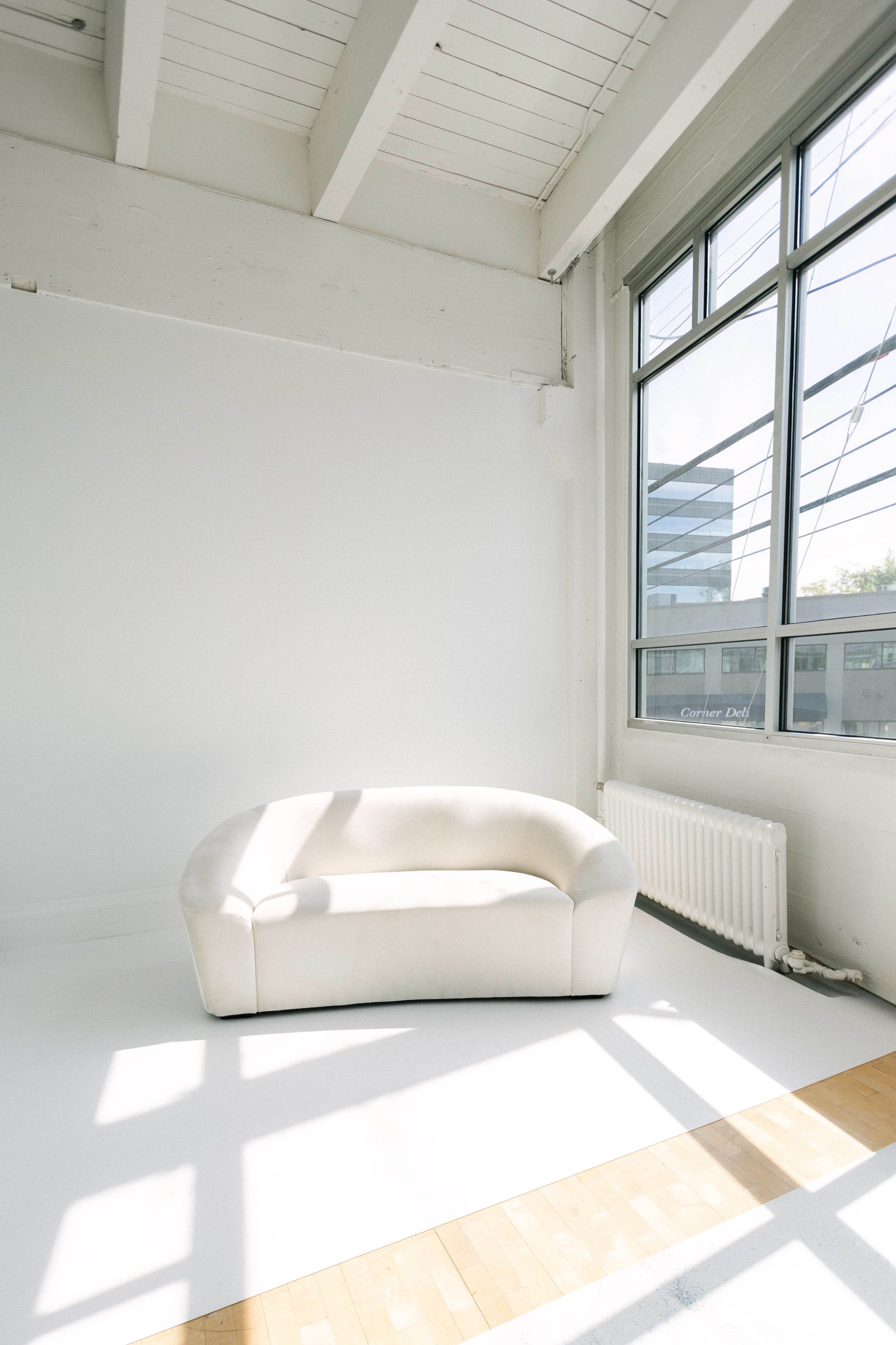 The image shows a white, curved sofa placed in a bright, minimalist room with large windows allowing natural light to enter.