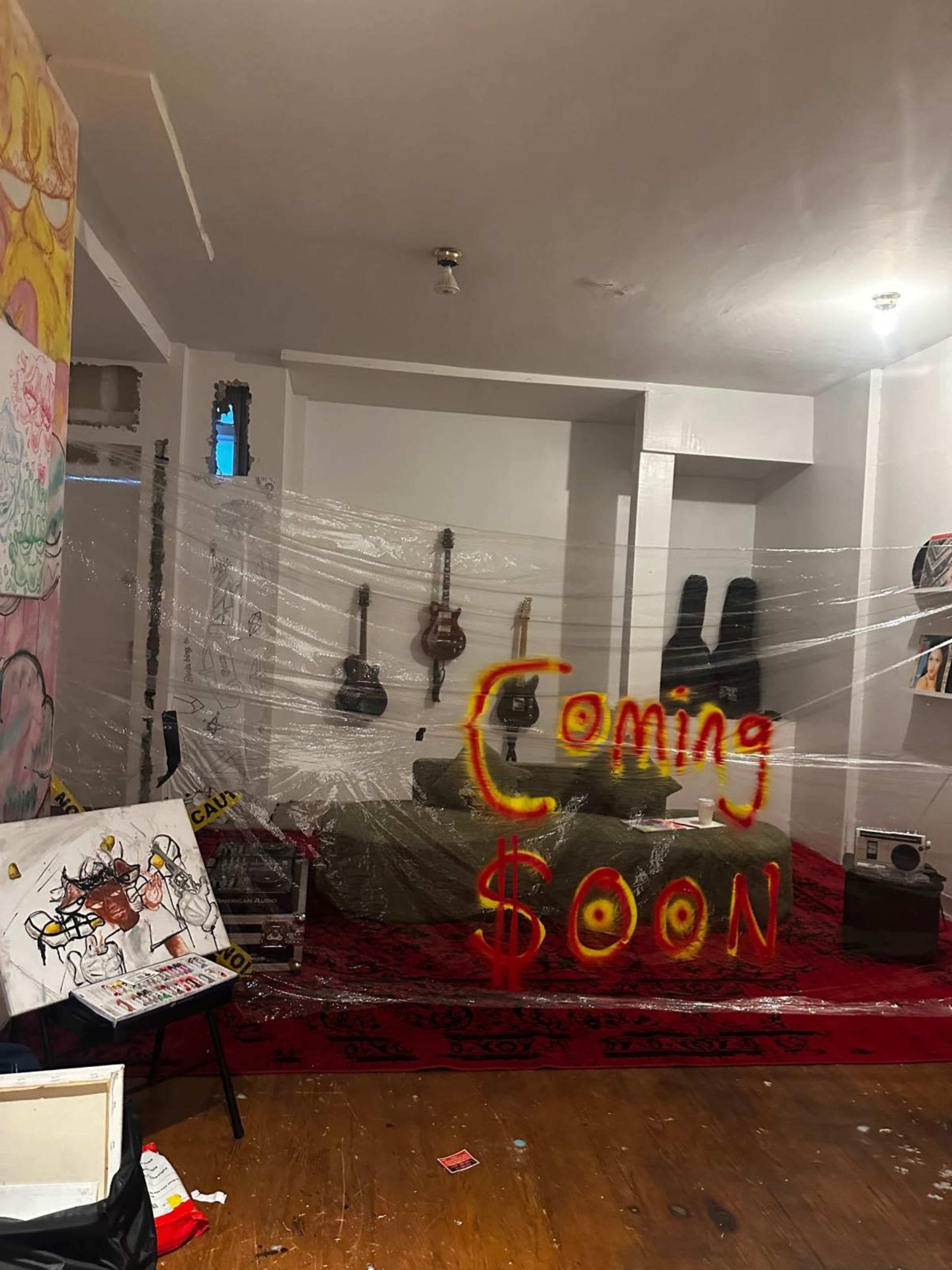 A room is partially enclosed with plastic wrap, featuring a red rug, guitars, and artwork, with the words "Coming Soon" displayed in yellow and red.