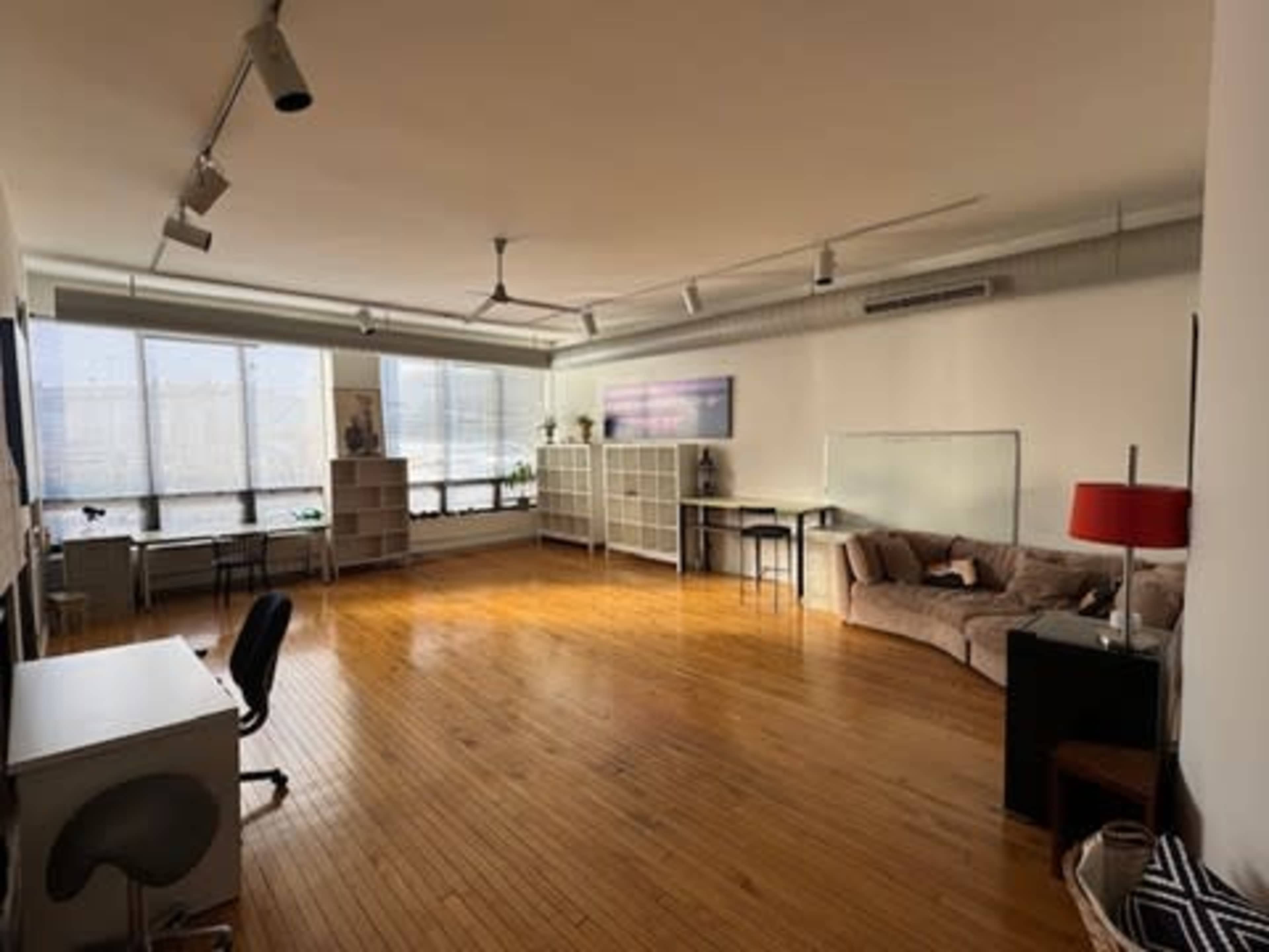 The image shows a spacious, bright room with wooden floors, large windows, and minimal furniture, including a couch, shelving units, and desks.