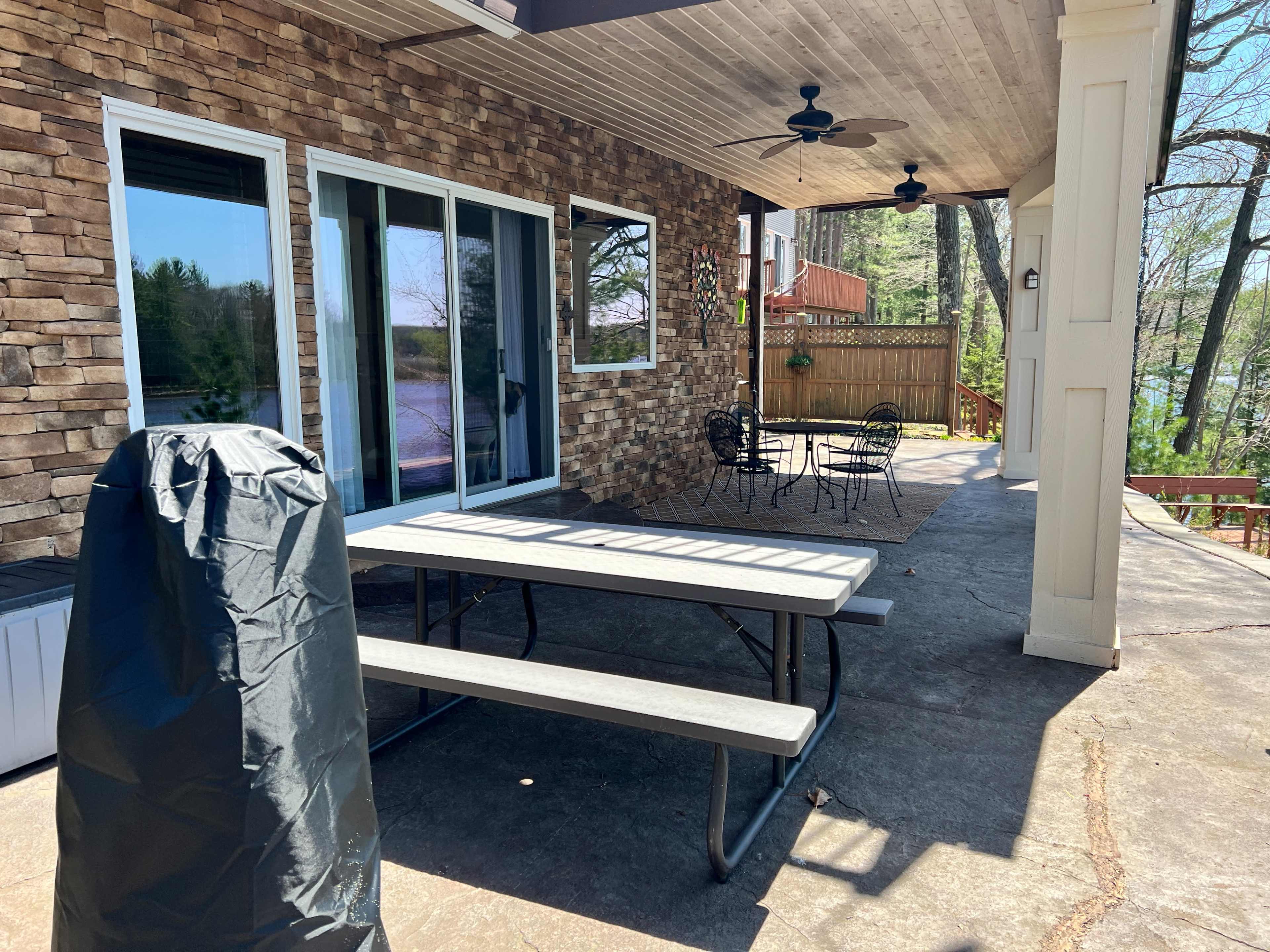 A covered patio features a picnic table, black outdoor chairs, and a charcoal grill cover, backed by stone walls and surrounded by trees.