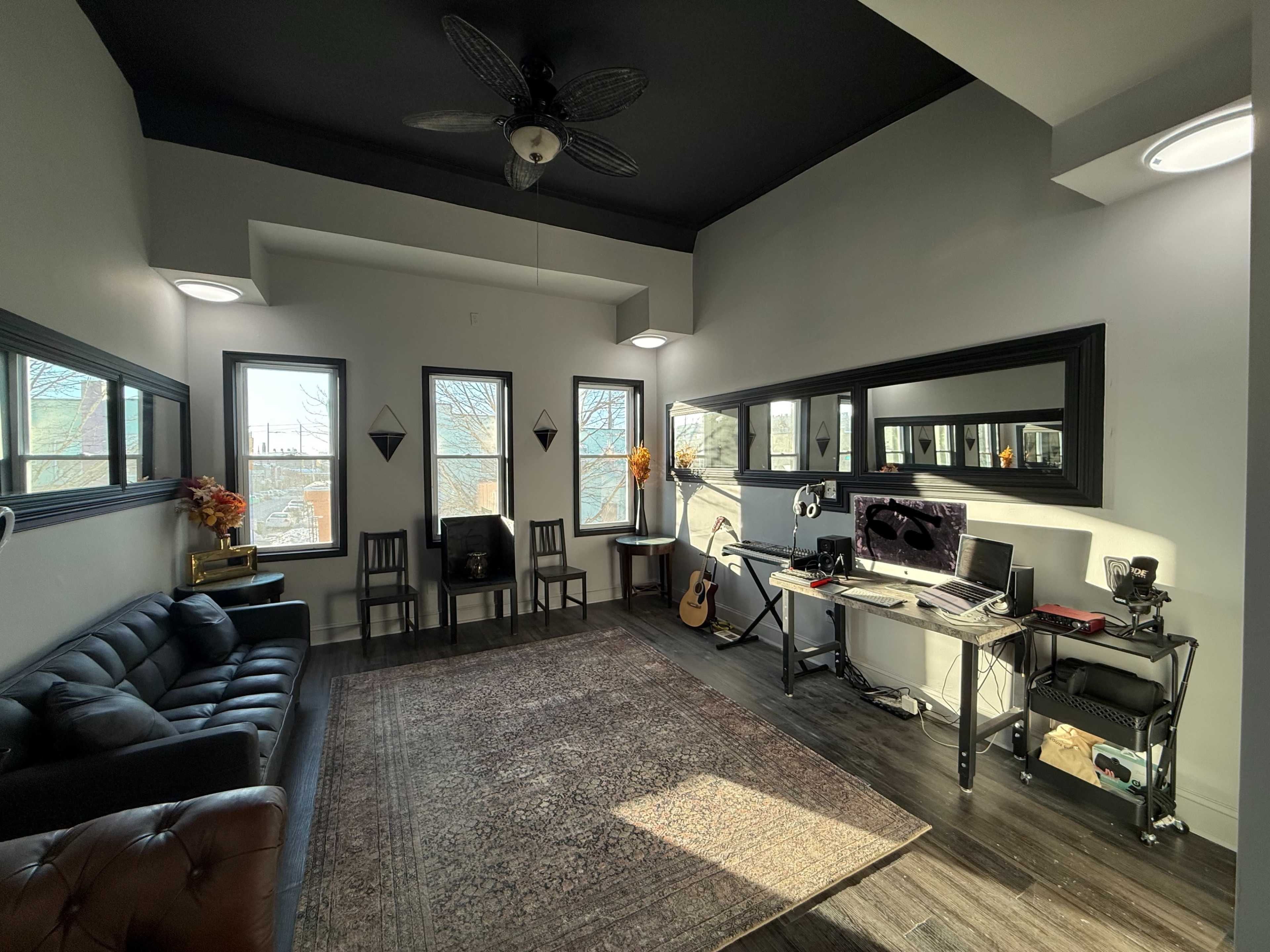 The image shows a modern living space with a black ceiling, a dark leather sofa, two chairs, a rug, and a desk equipped with a computer and musical equipment.