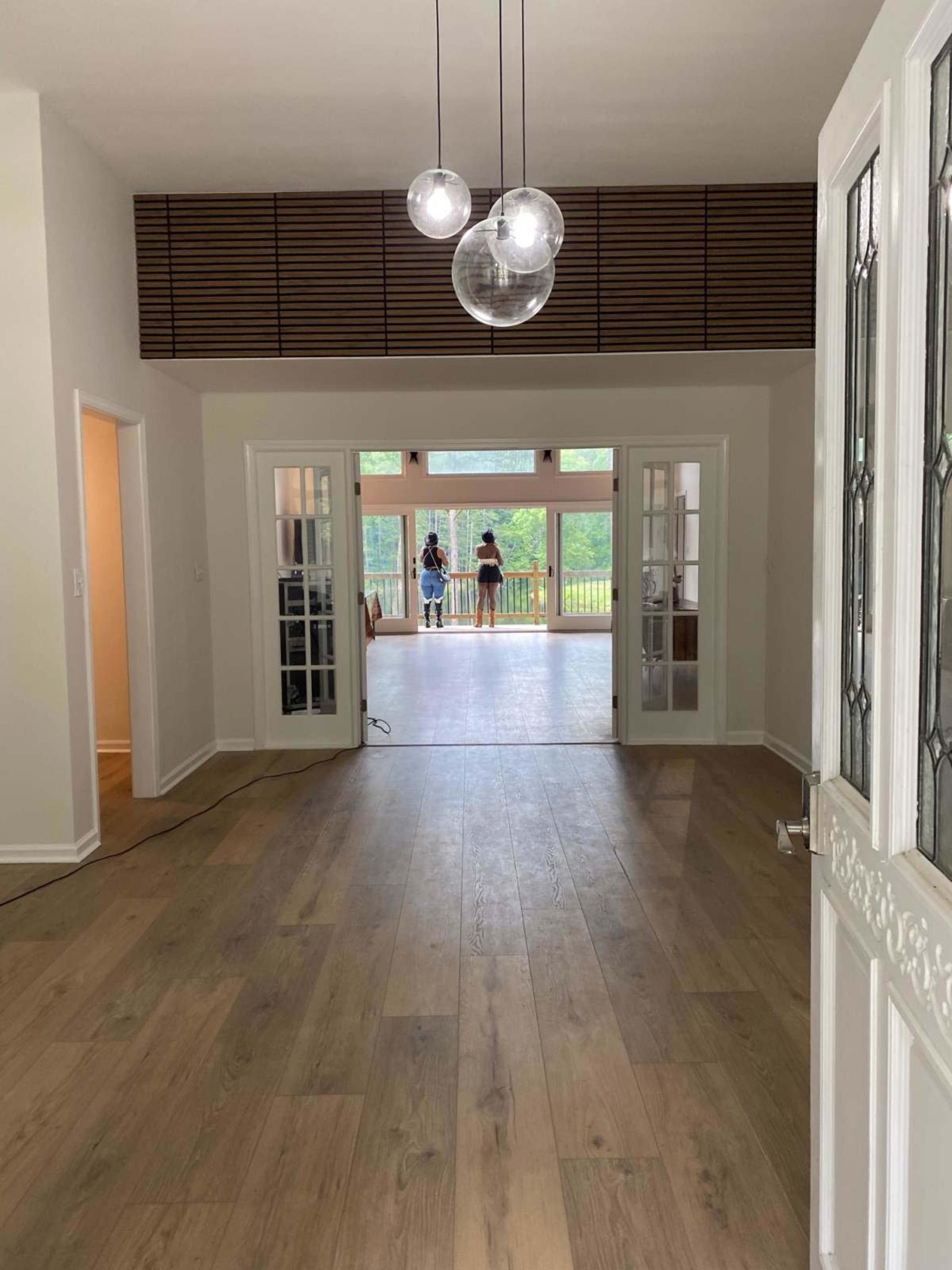 The image shows an interior view of a room with wooden flooring, glass-paneled doors, and a large opening leading to a deck area with two people standing outside.