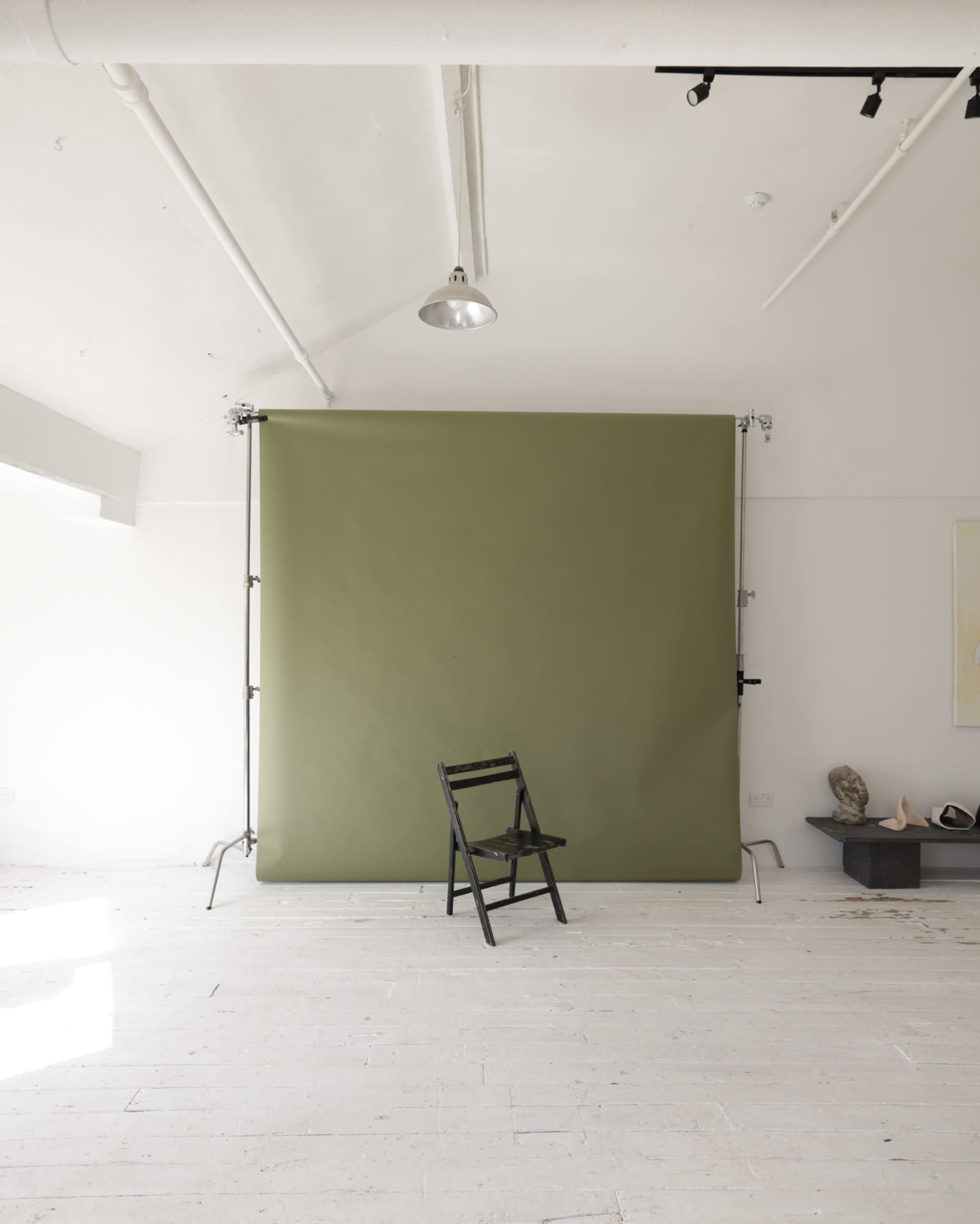 The image shows a minimalist studio space with a green backdrop and a black folding chair positioned in front of it.
