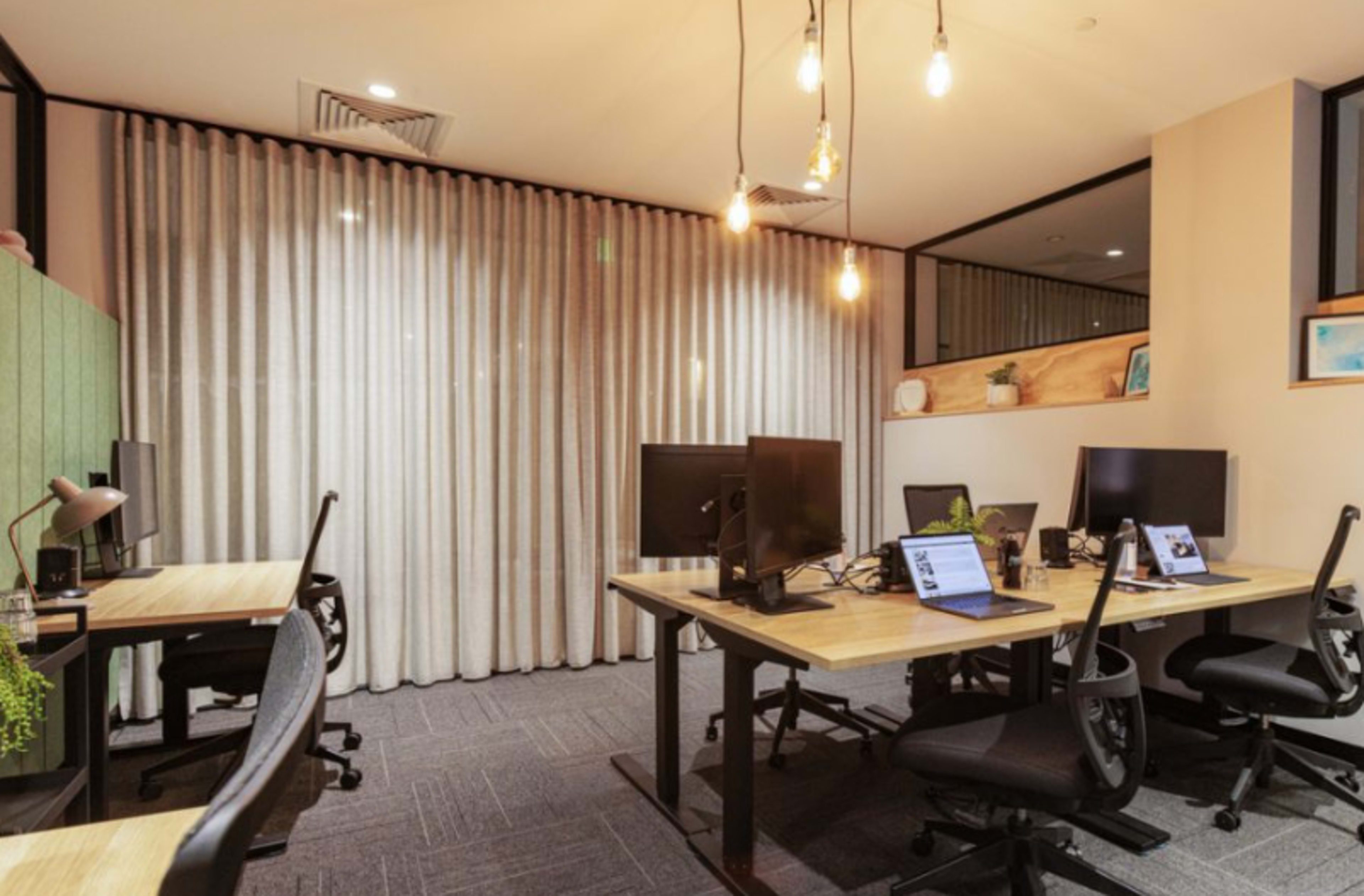 A modern office space featuring several desks with computer monitors, hanging light fixtures, and a wall of curtains.