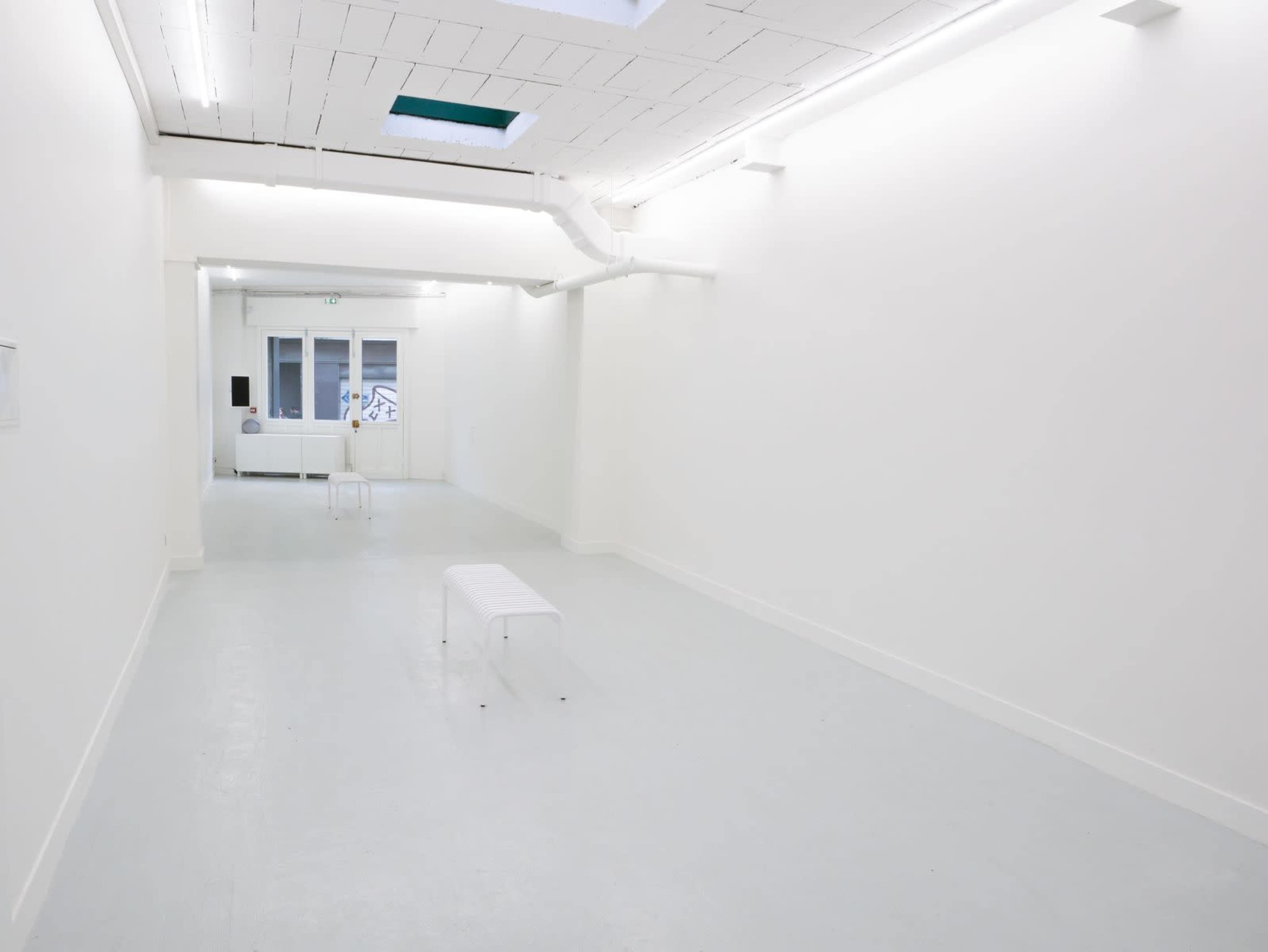 The image shows a minimalist gallery space with white walls, a light gray floor, and a few transparent benches.
