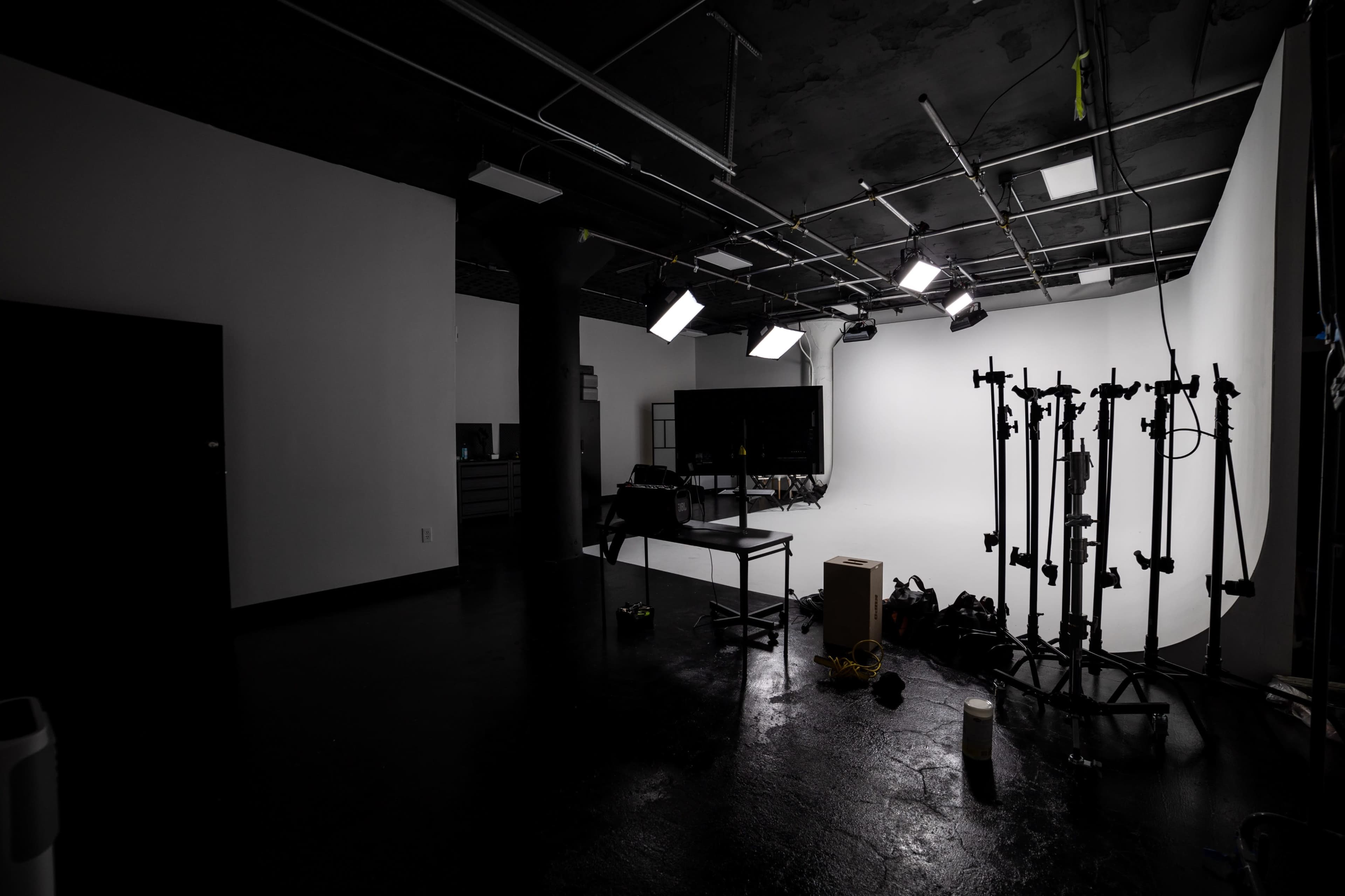 A dimly lit studio space features a blank white backdrop, several lighting fixtures, and equipment stands arranged near a black seating area.